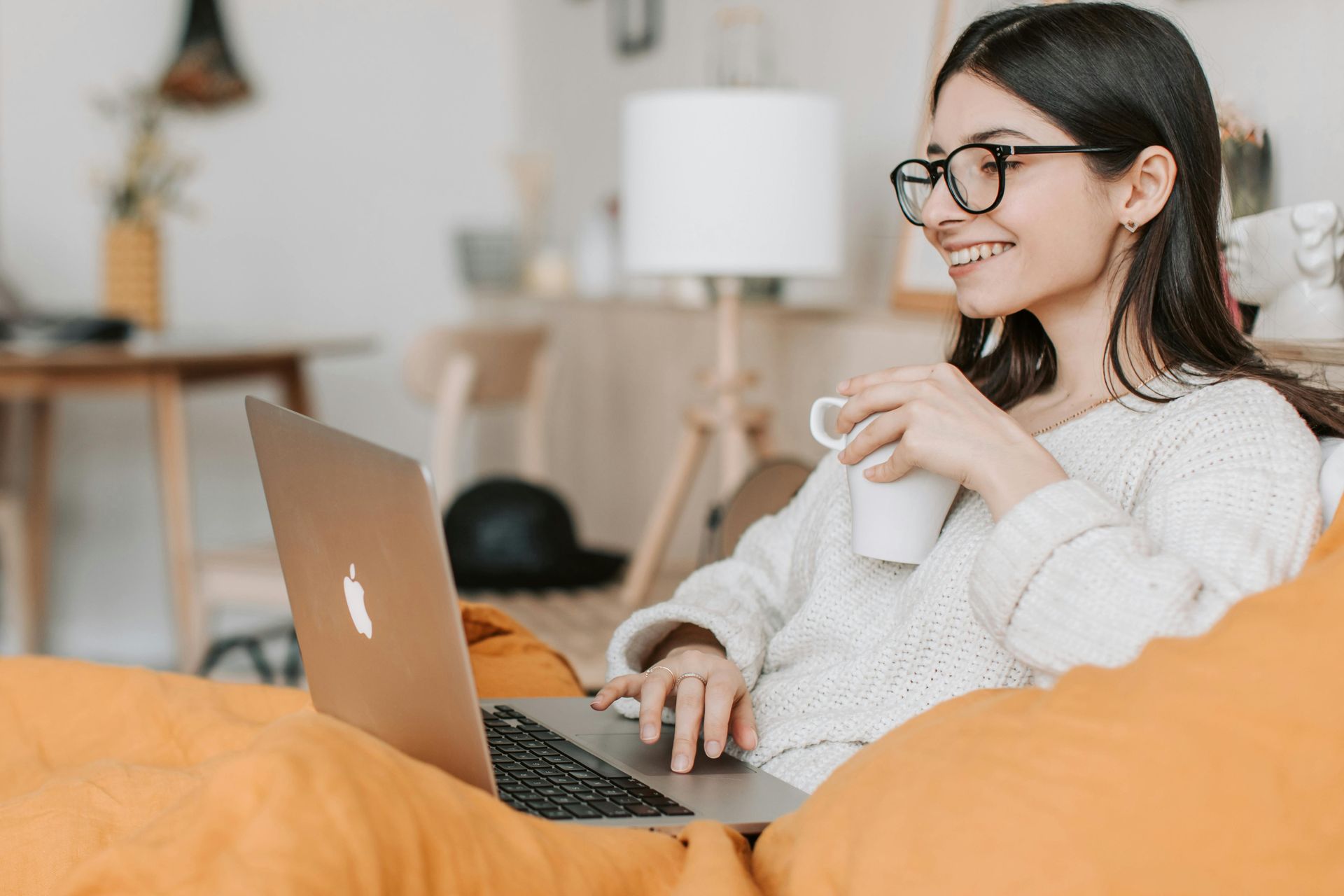 A woman is sitting on a bean bag chair using a laptop computer and drinking coffee.