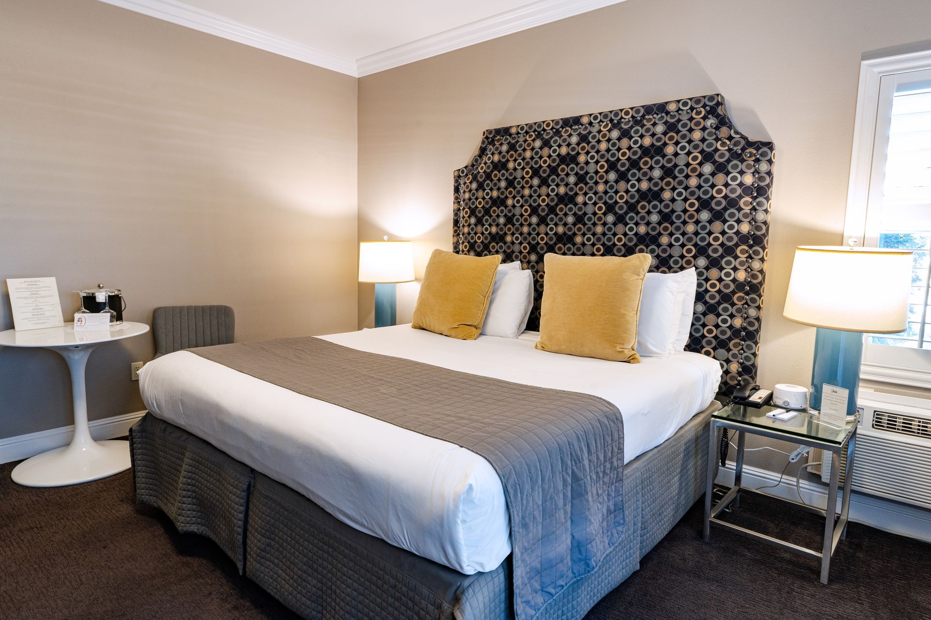 Hotel room with a large bed, decorative headboard, side table, lamps, and a small table with a chair.