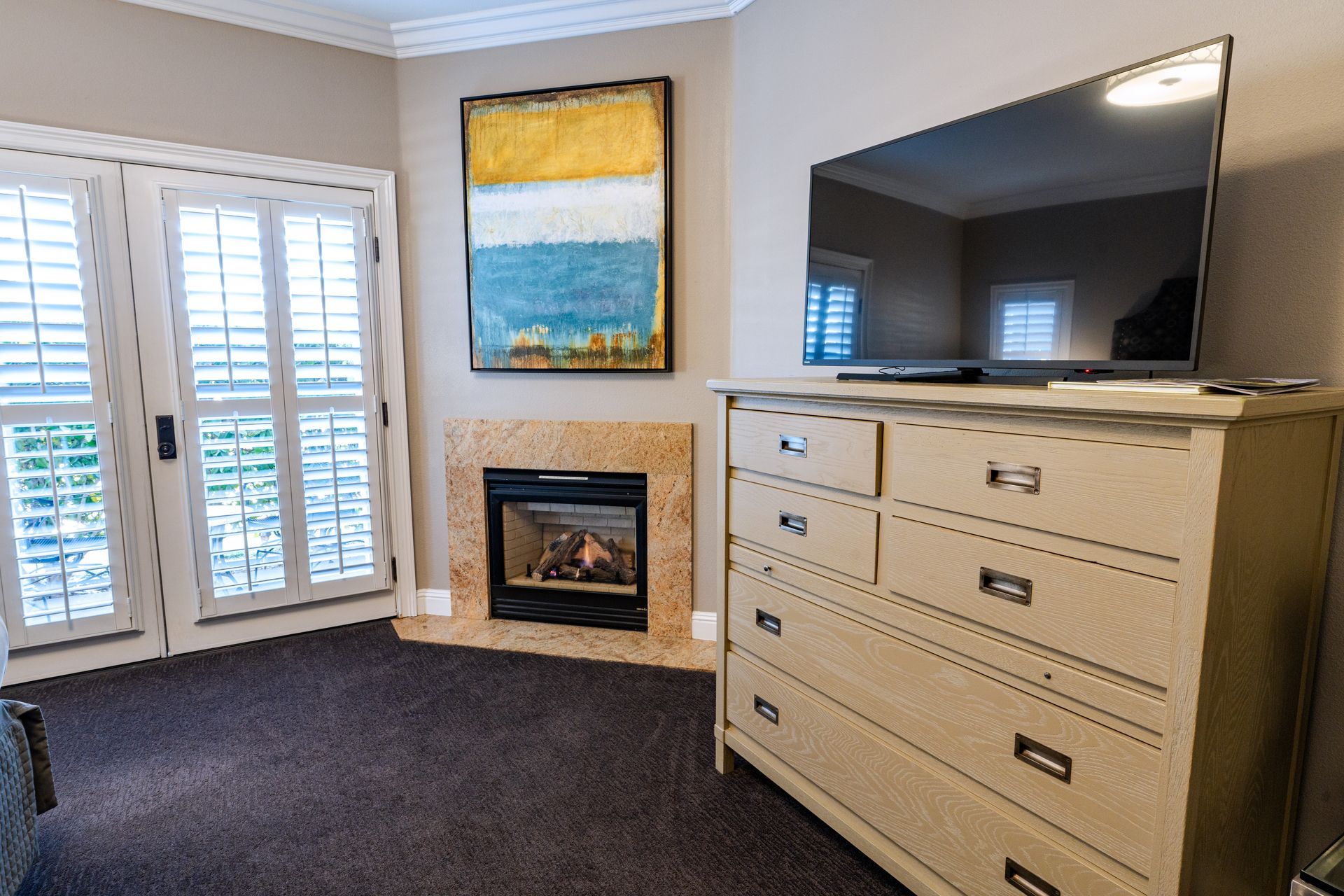 Bedroom with a fireplace, TV, and dresser. French doors with shutters and a colorful painting hang on the wall.