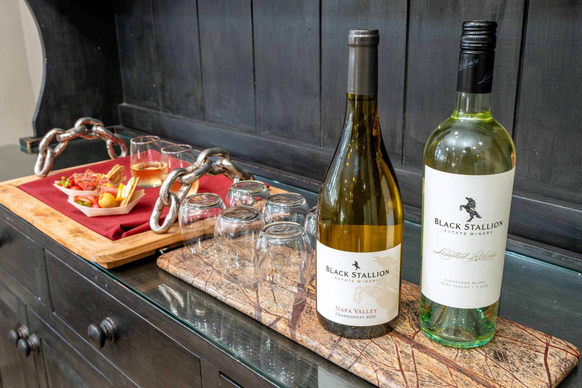 Two bottles of white wine, glasses, and snacks on a wood serving board atop a dark cabinet.