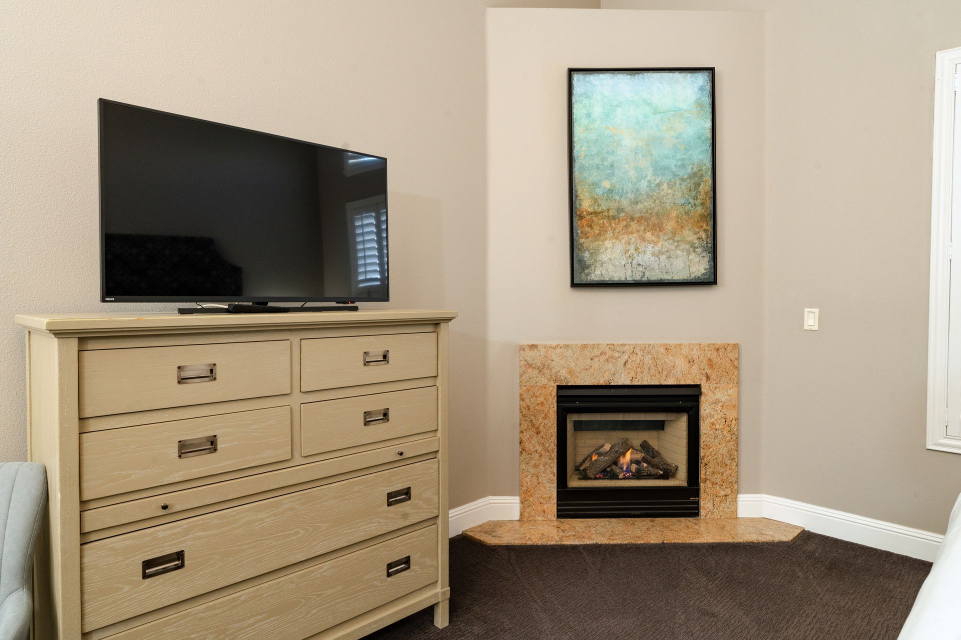 Bedroom with TV on dresser, art and fireplace. Neutral walls, brown carpet.