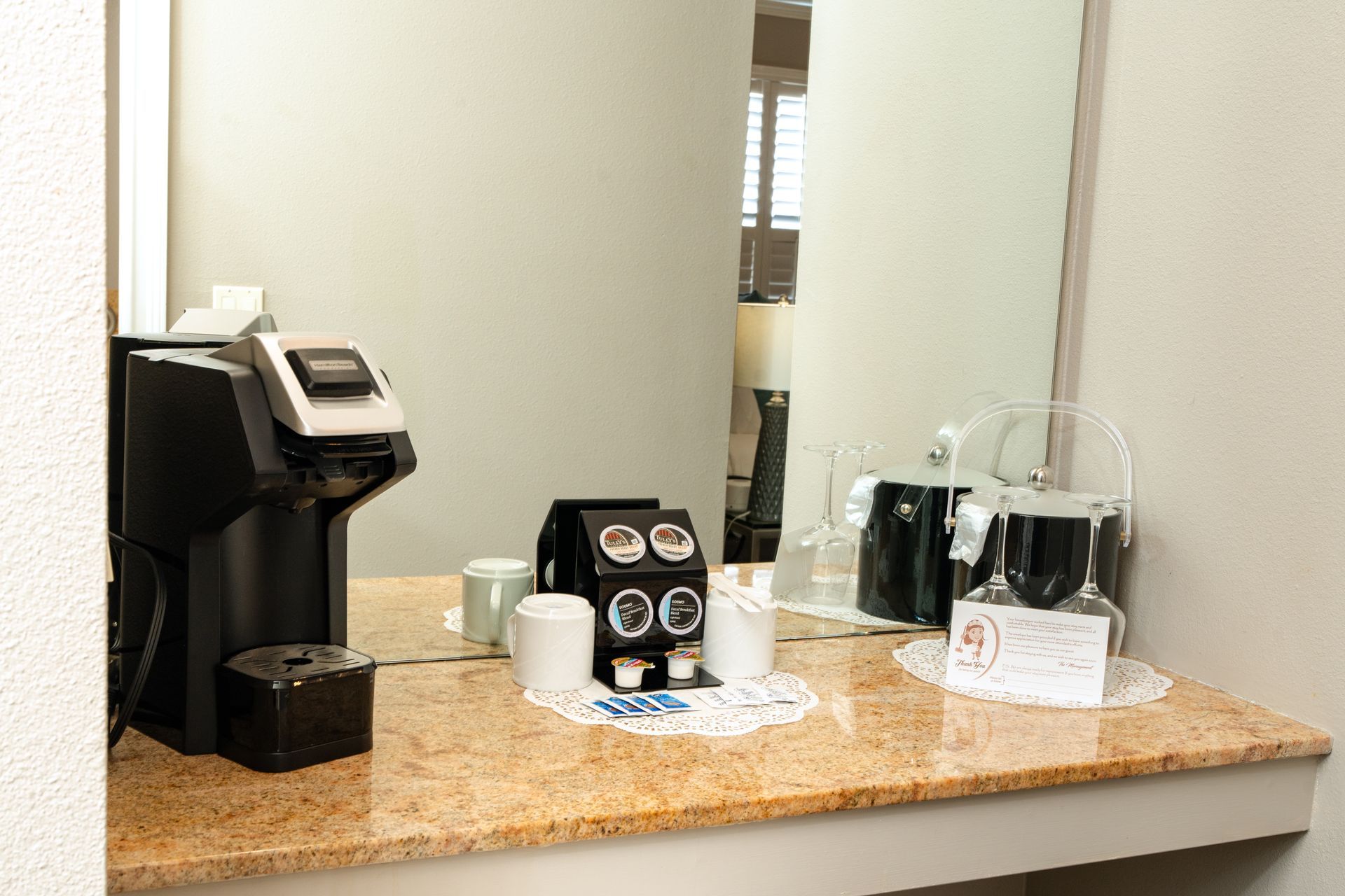 Coffee station on a countertop with a Keurig, coffee pods, cups, a carafe, and a mirror.