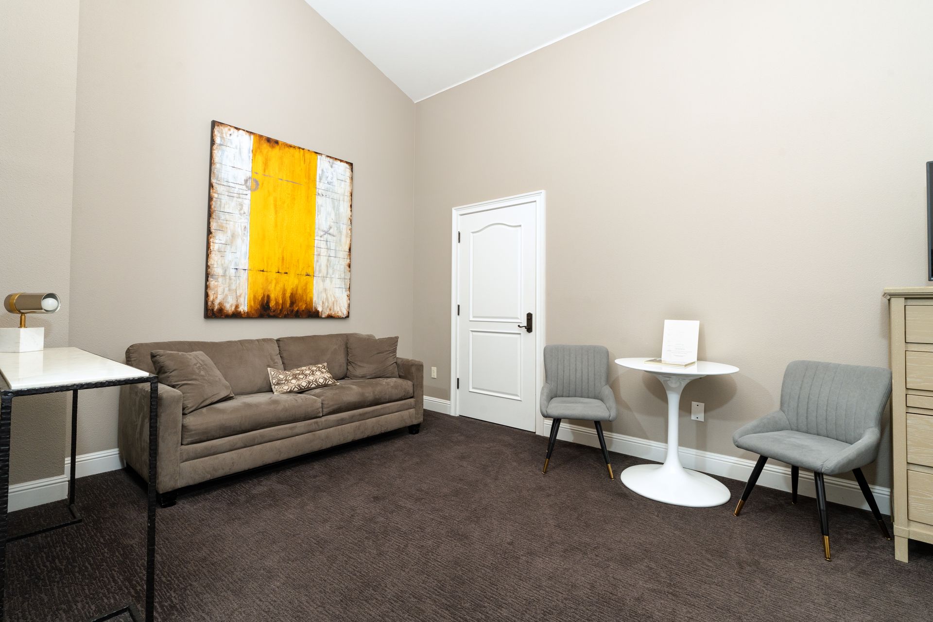 Neutral-toned room with brown couch, white table, two gray chairs, and large artwork. Dark brown carpet.