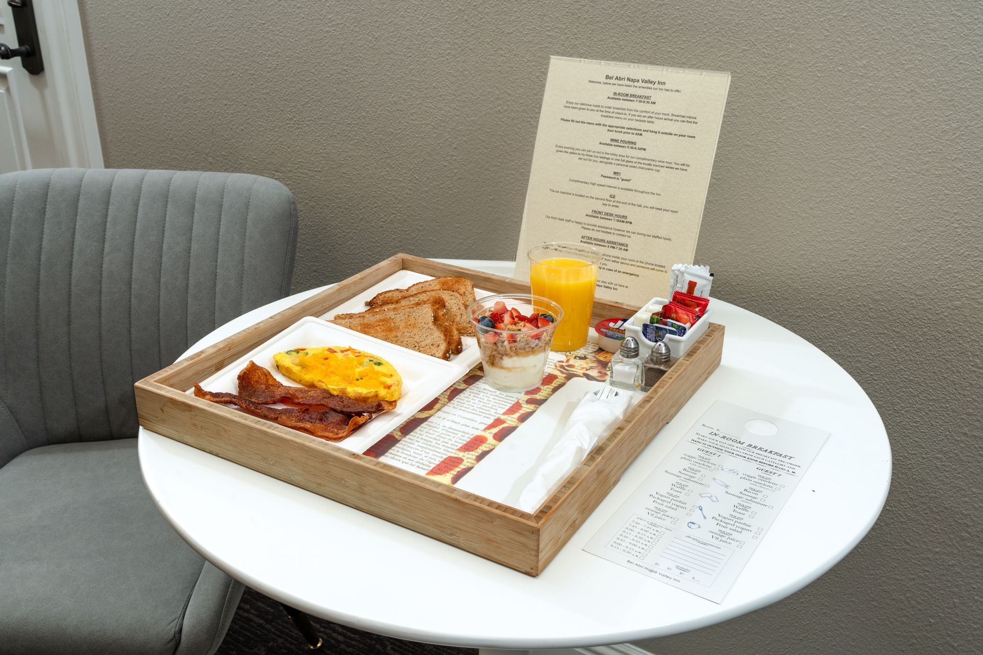 Breakfast tray on a small white table, including toast, fruit, juice, and condiments, next to a chair and menu.