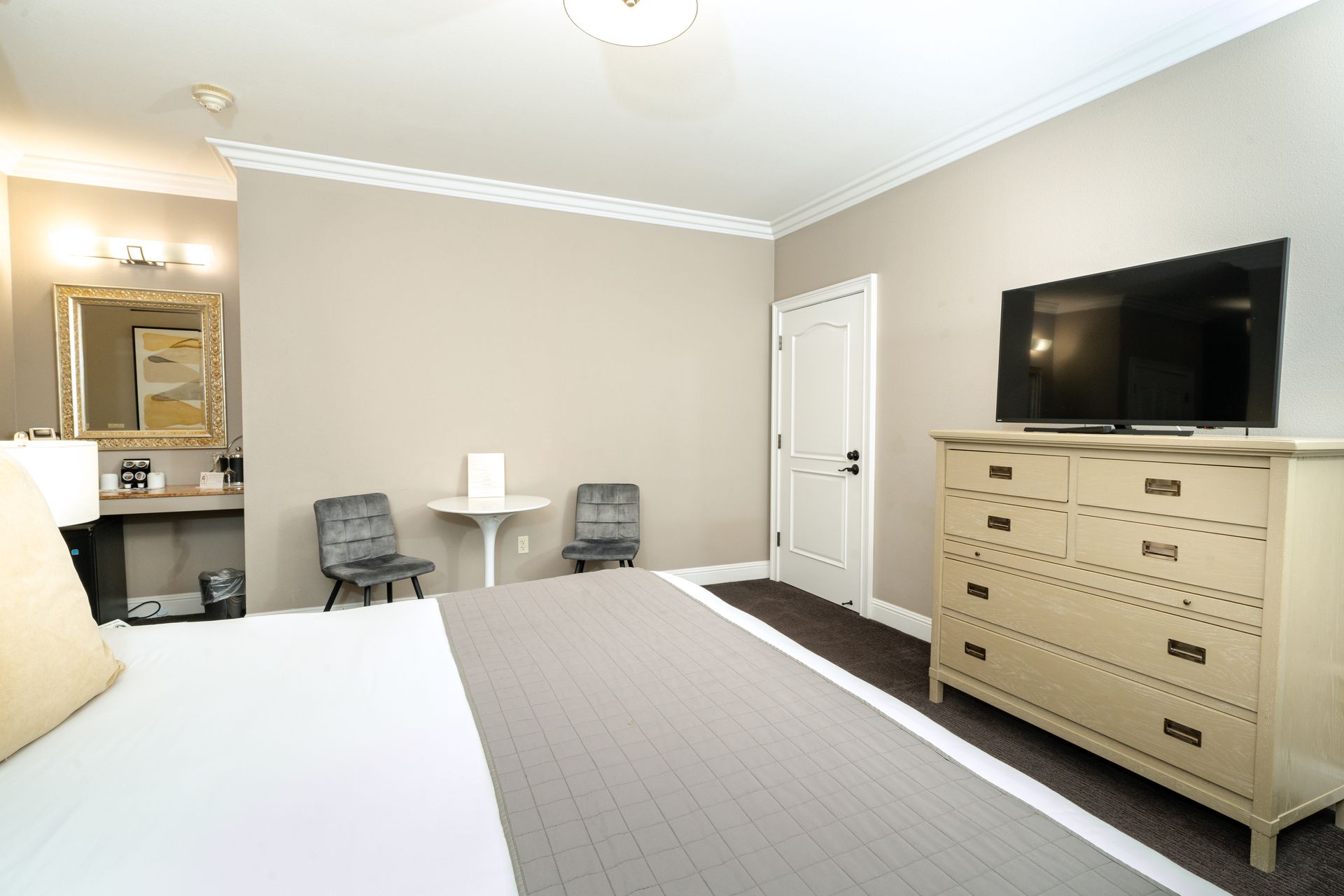 Bedroom with a bed, dresser, table, chairs, TV, and vanity. Beige and gray color scheme.