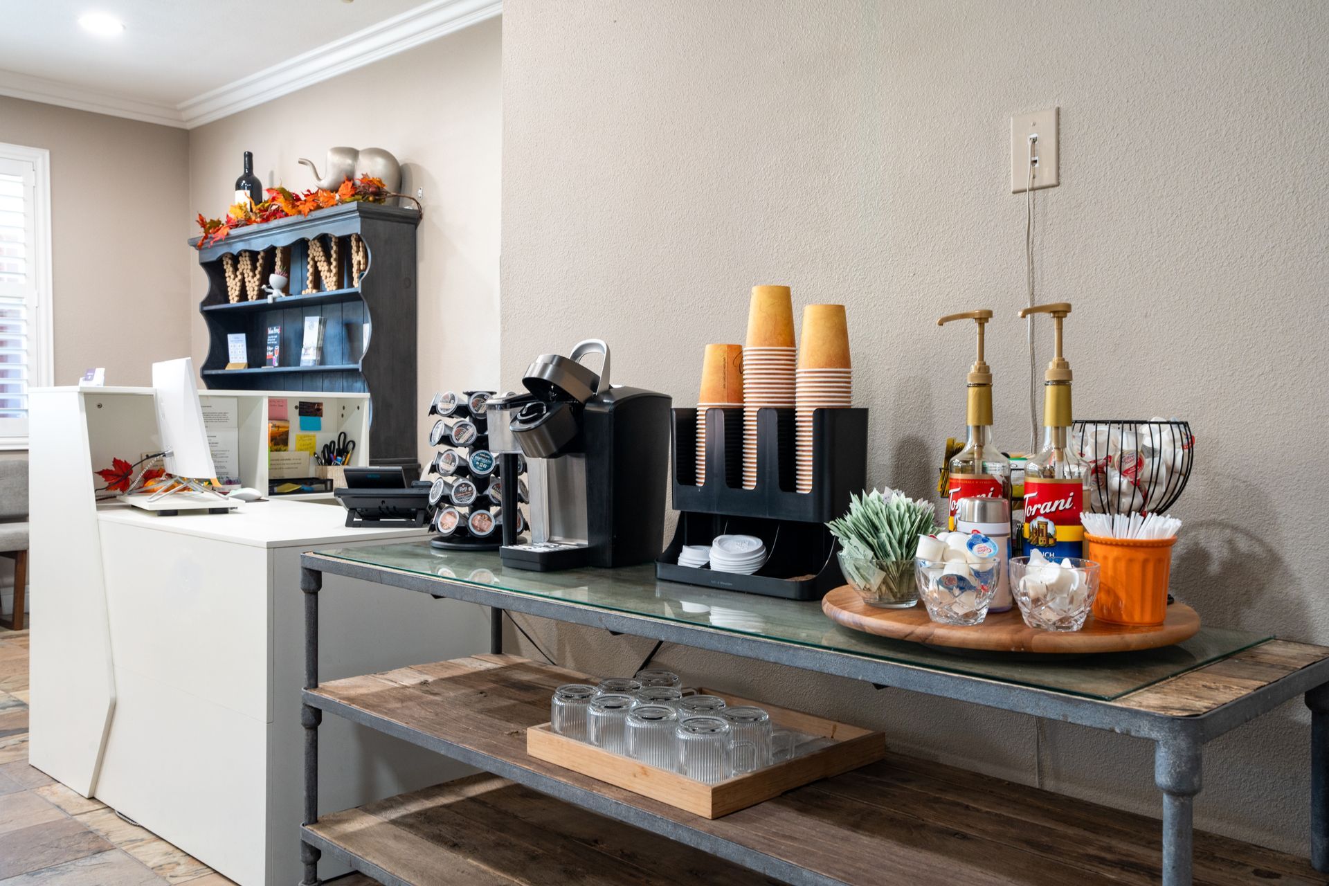 Coffee station on a table with a Keurig, cups, syrups, and other accessories in a well-lit office.