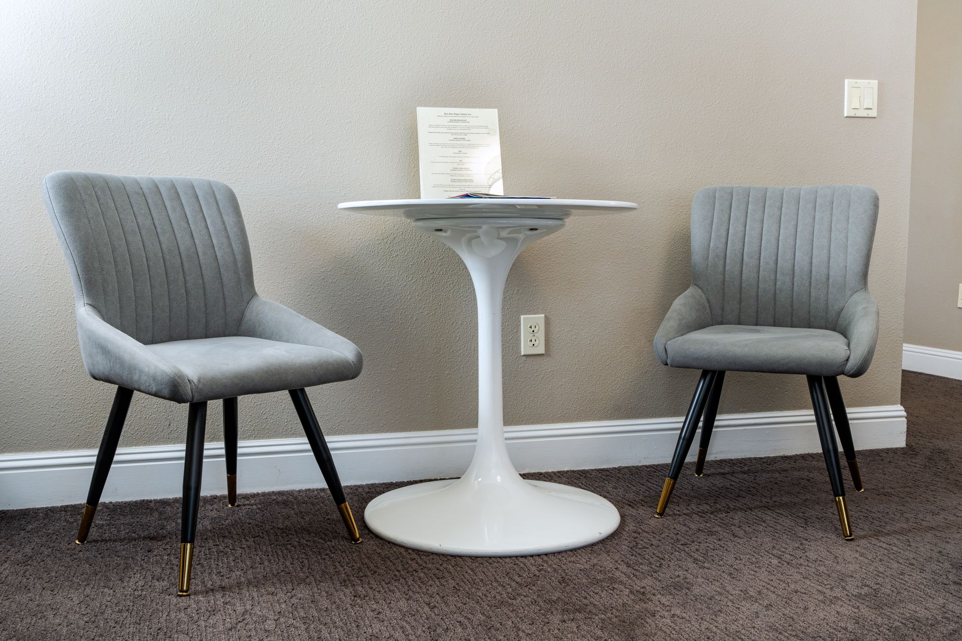 Two gray chairs face a white tulip table in a room with light brown walls and dark carpet.