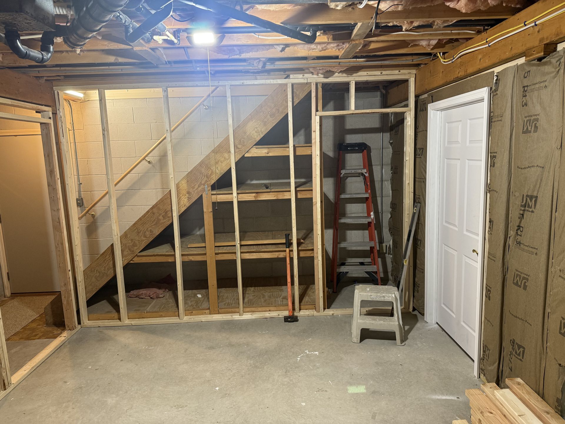Unfinished basement with exposed framing, staircase, ladder, and a white door under construction.