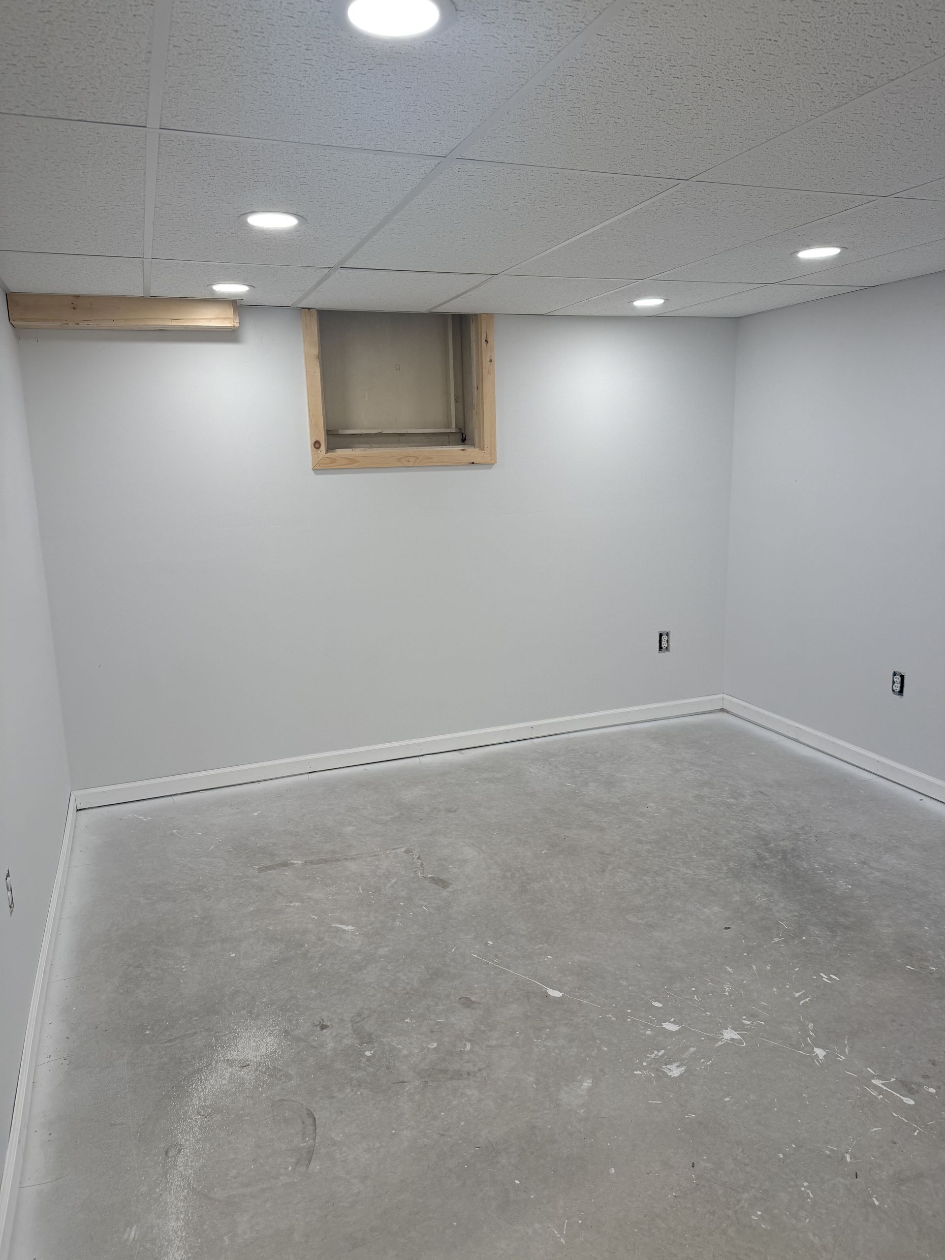 Empty basement room with white walls, drop ceiling, small window, and unfinished concrete floor
