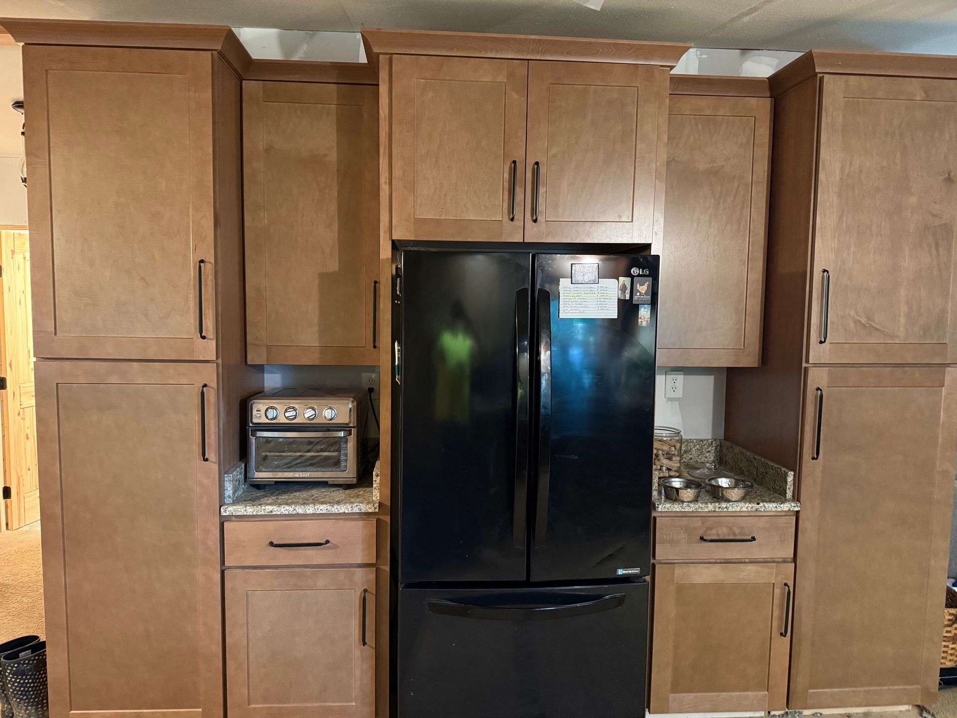 Wooden kitchen cabinets around a black refrigerator and toaster oven, with granite countertops.