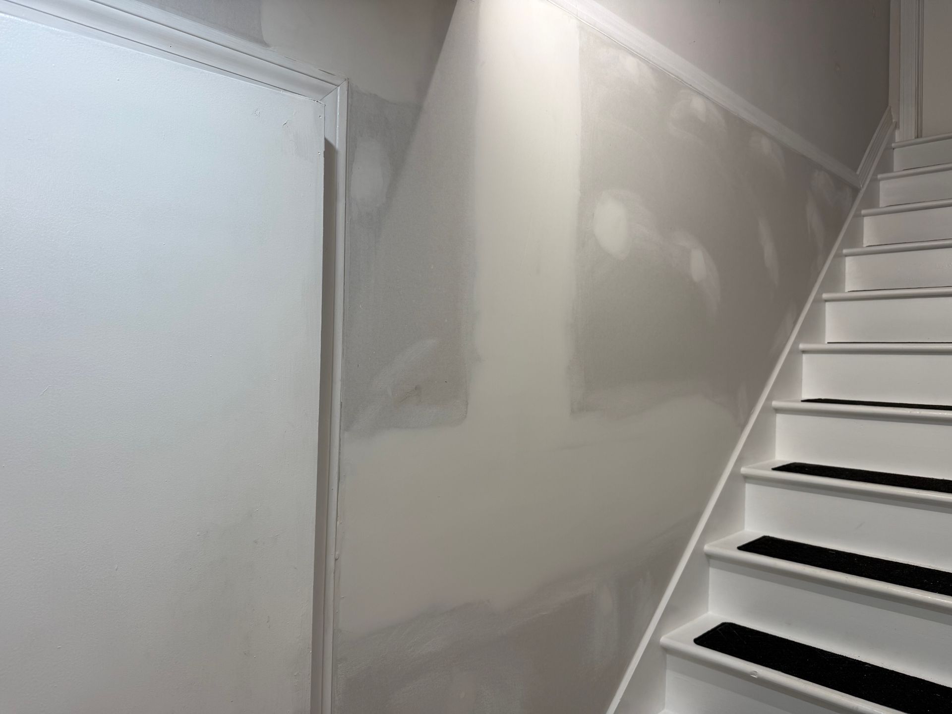Narrow stairwell with white walls and stairs under renovation, showing patchy drywall and unfinished surfaces