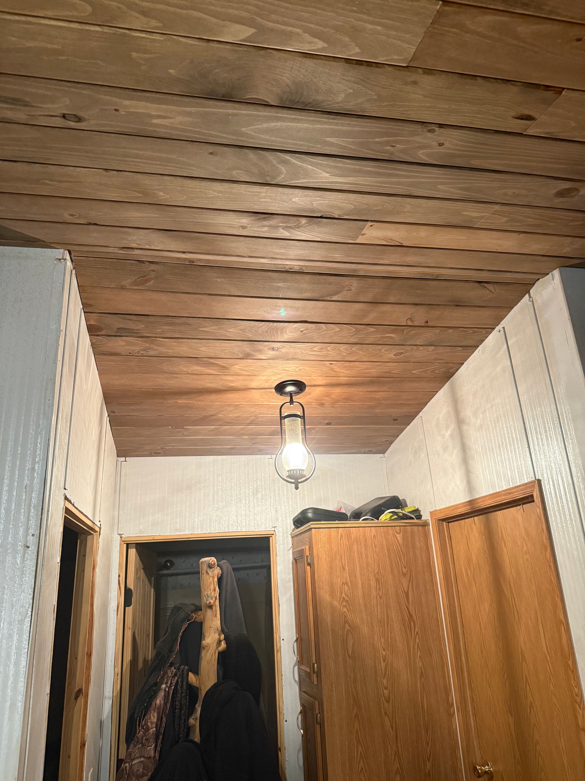 Narrow hallway with wood-paneled ceiling, hanging light, and coat racks by open doorways