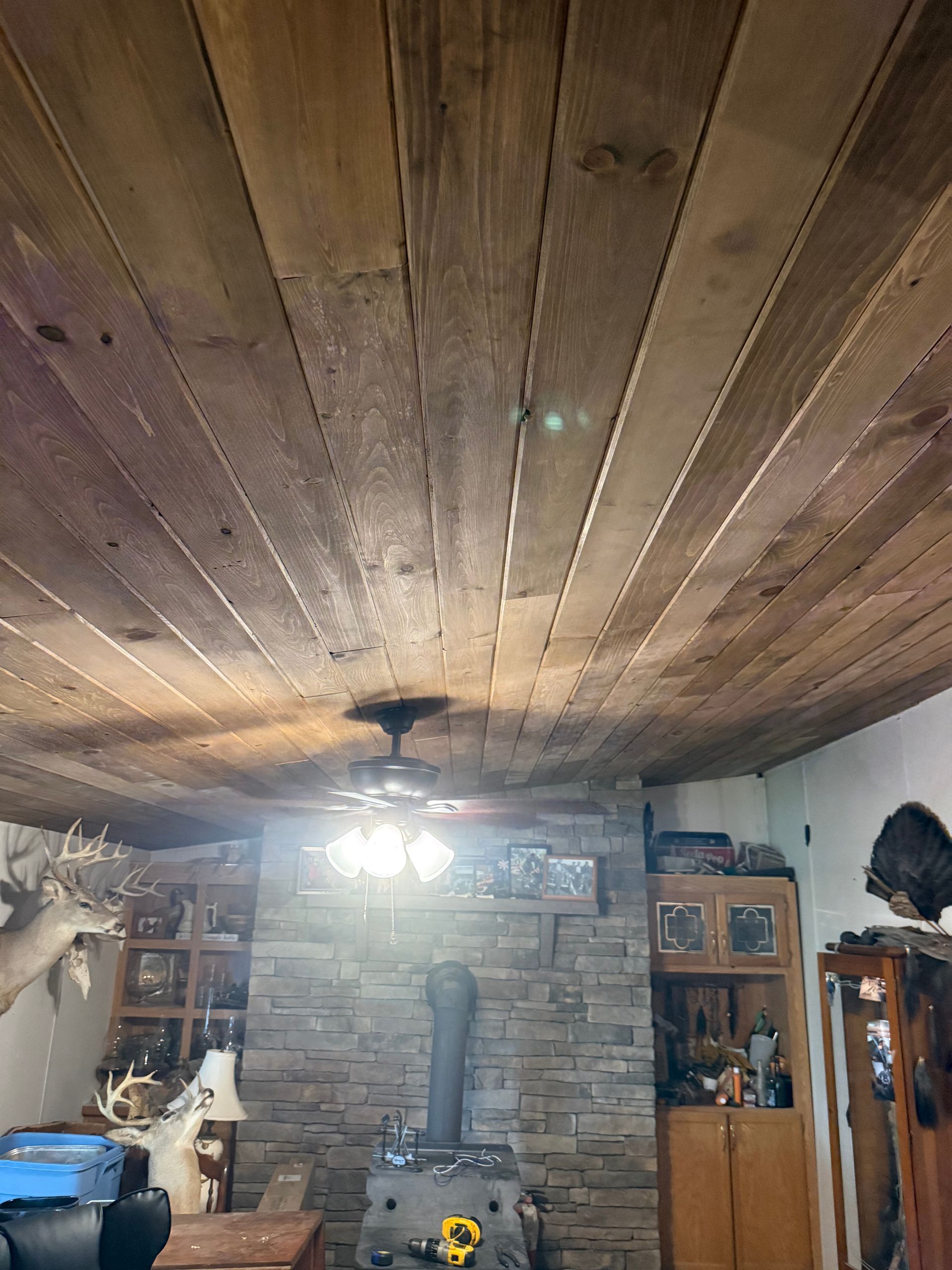 Wood-paneled ceiling over a stone fireplace in a rustic living room with shelves and a hanging light fixture