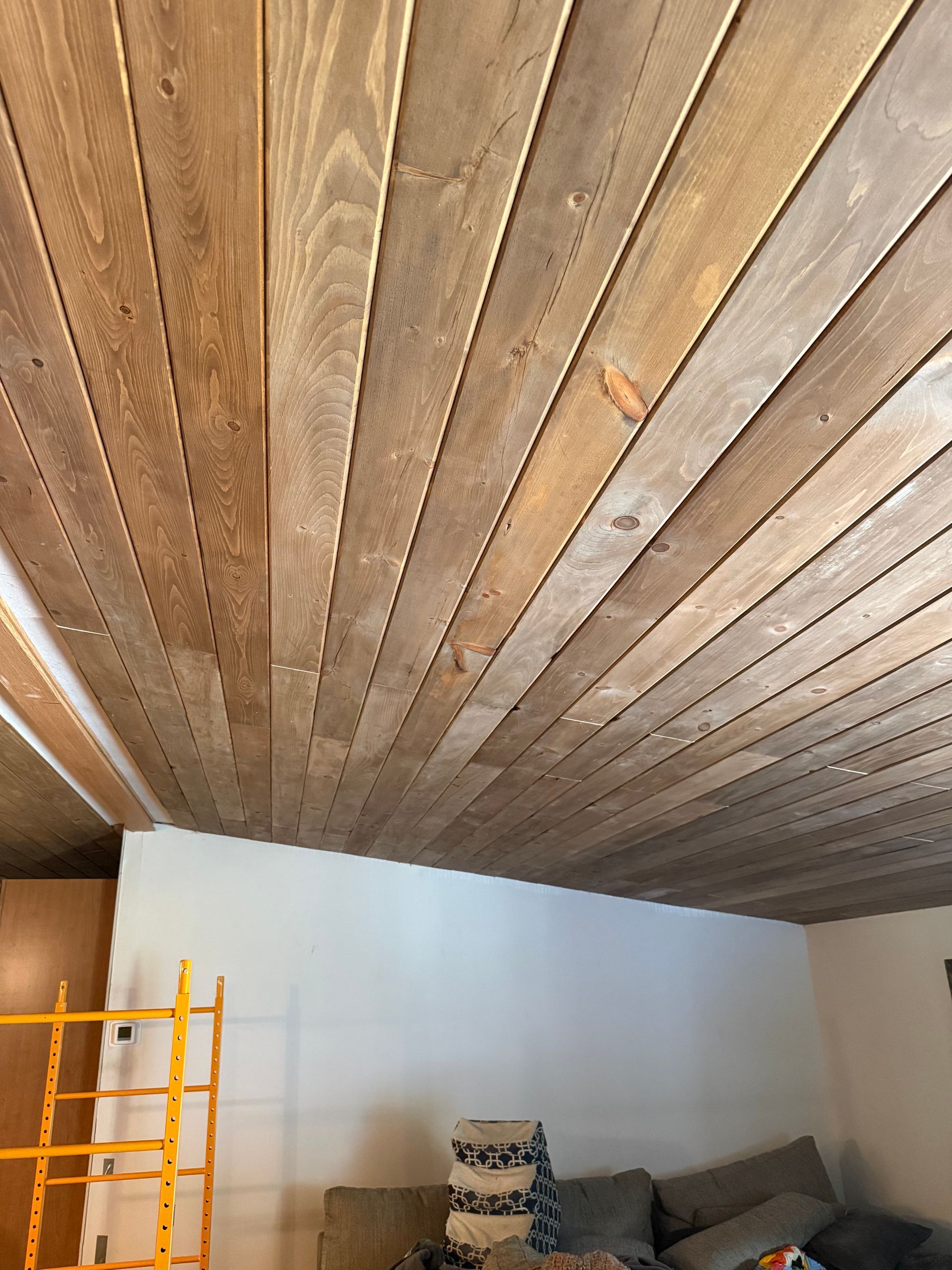 Wood plank ceiling over a living room with white walls, gray sofa, and yellow ladder on the left