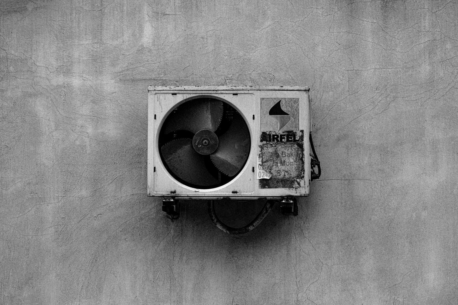 Wall-mounted air conditioner unit on a gray concrete wall, with a large fan visible on the left.