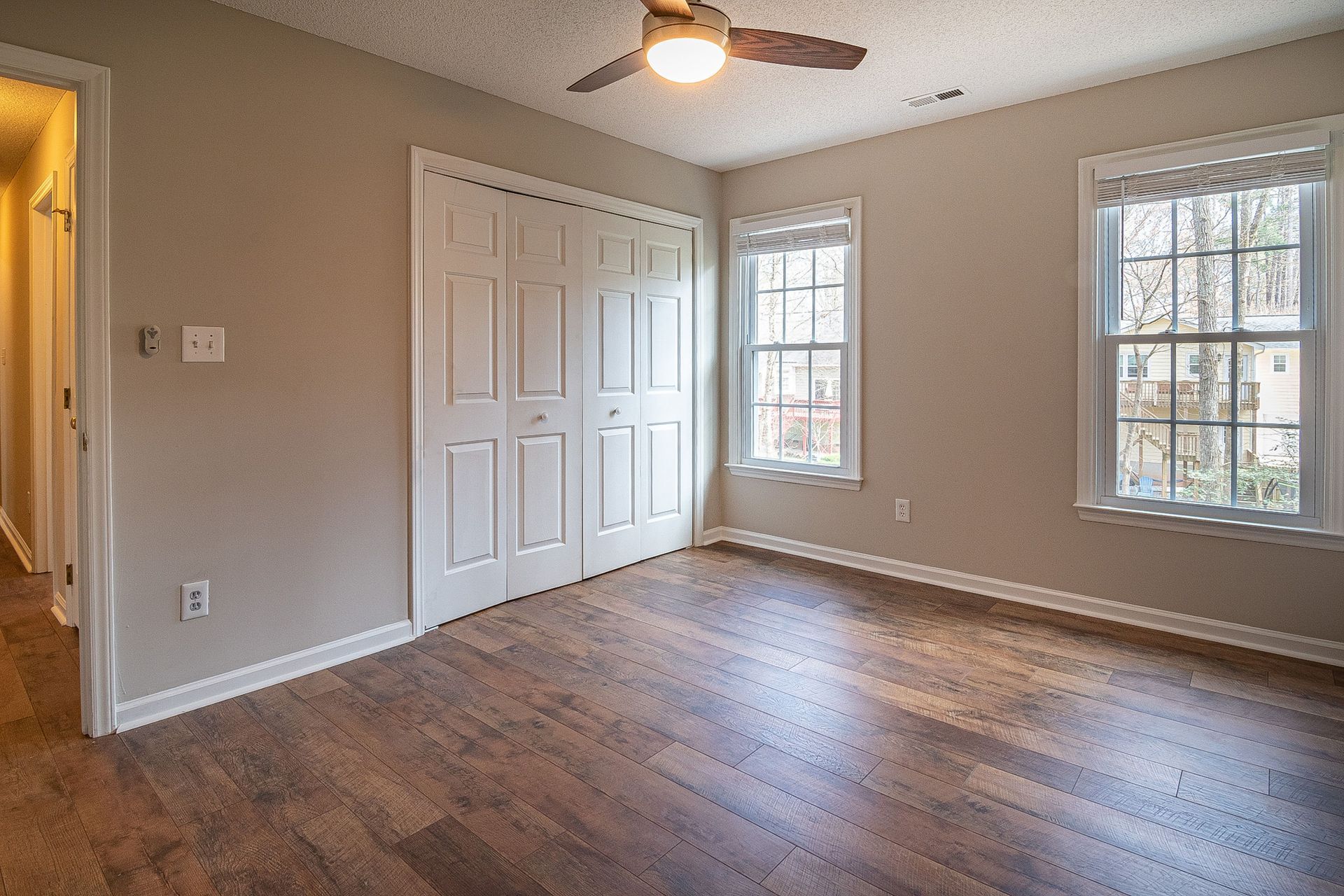 Empty beige bedroom with hardwood floors, ceiling fan, and two windows with white blinds.