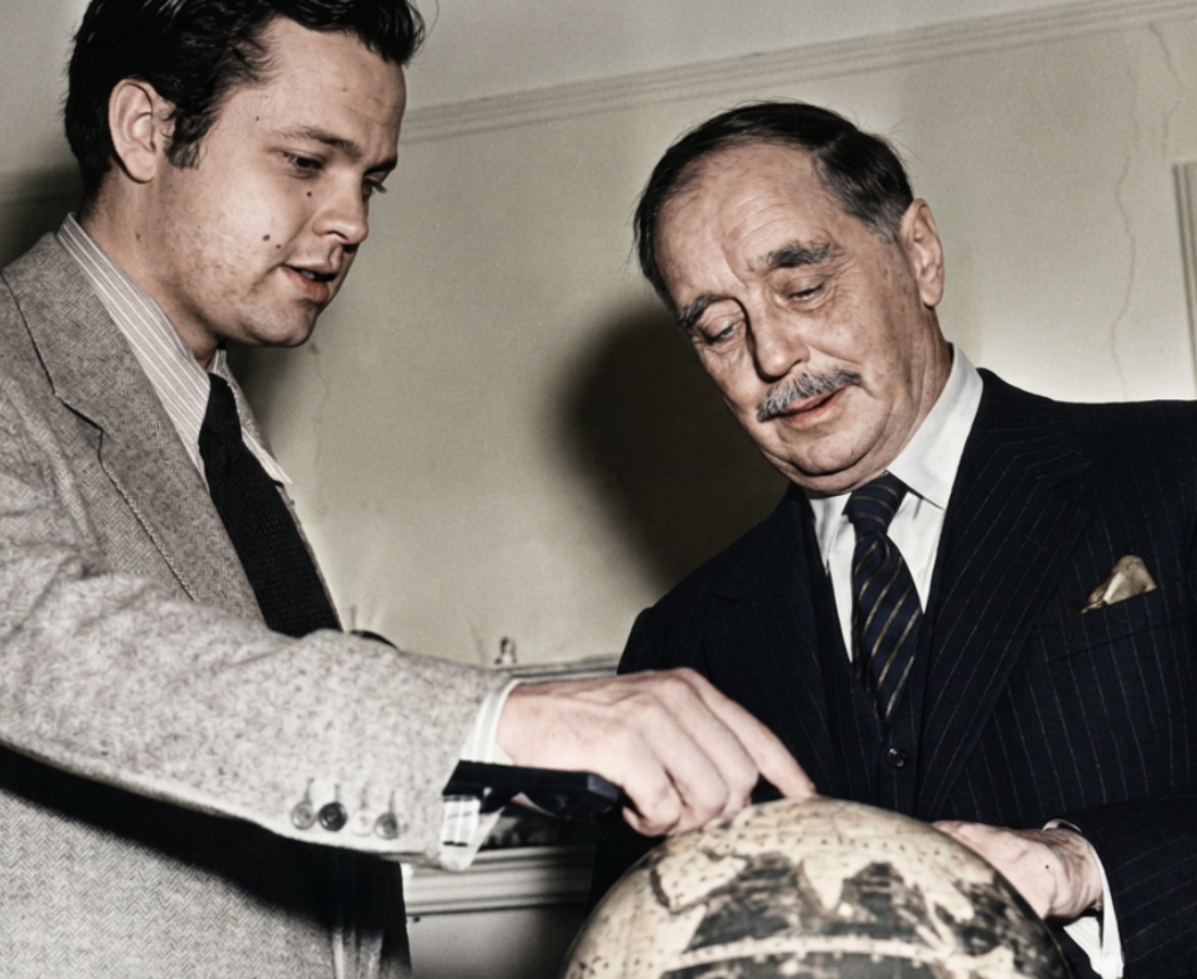 Black and white photo of H.G. Wells and the young Orson Welles meeting for a radio interview about the infamous War Of The Worlds 'panic broadcast' of 1938. dressed in formal suits, they are pointing at a globe of the world.