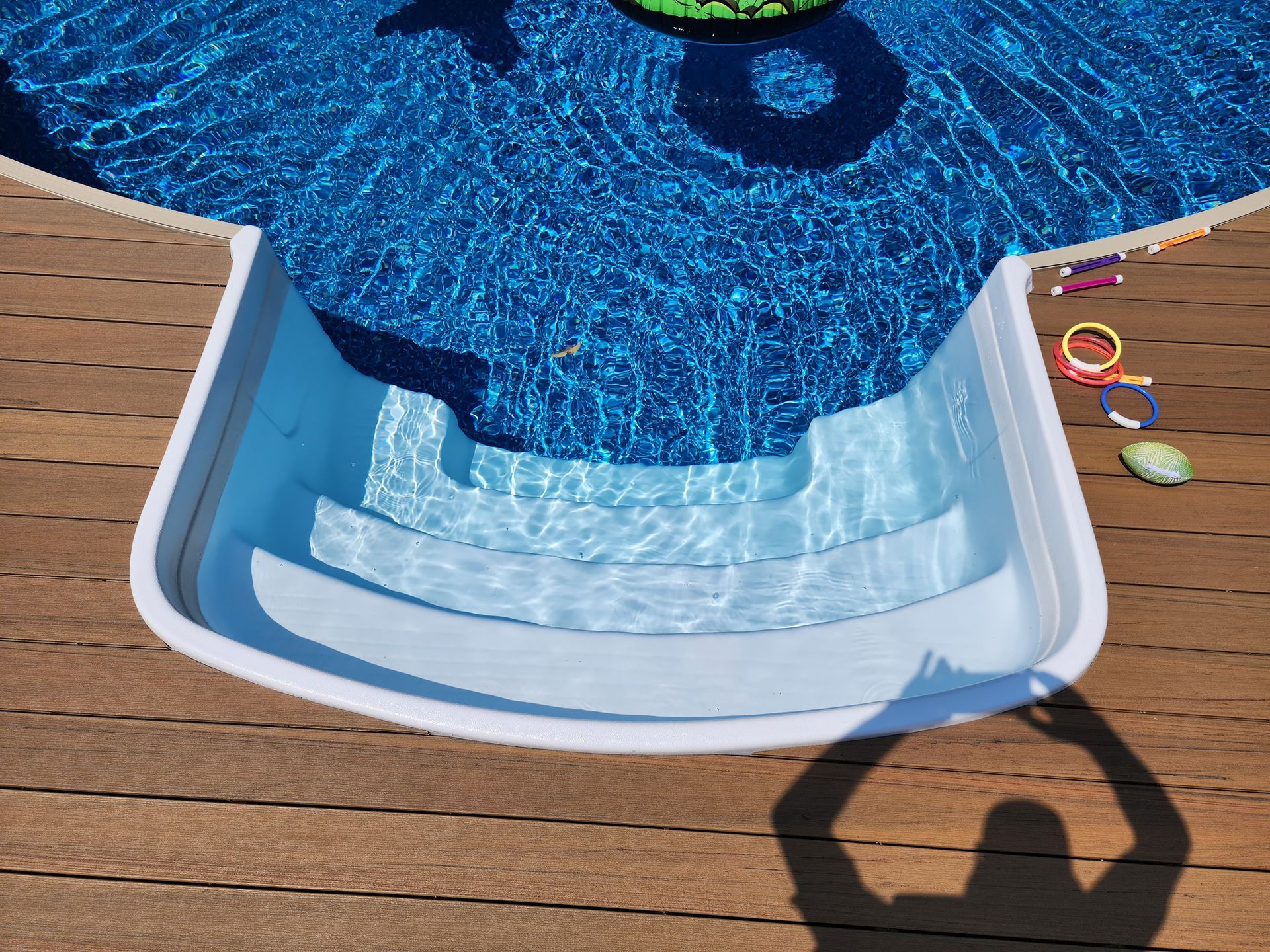 A shadow of a person is cast on a wooden deck next to a swimming pool