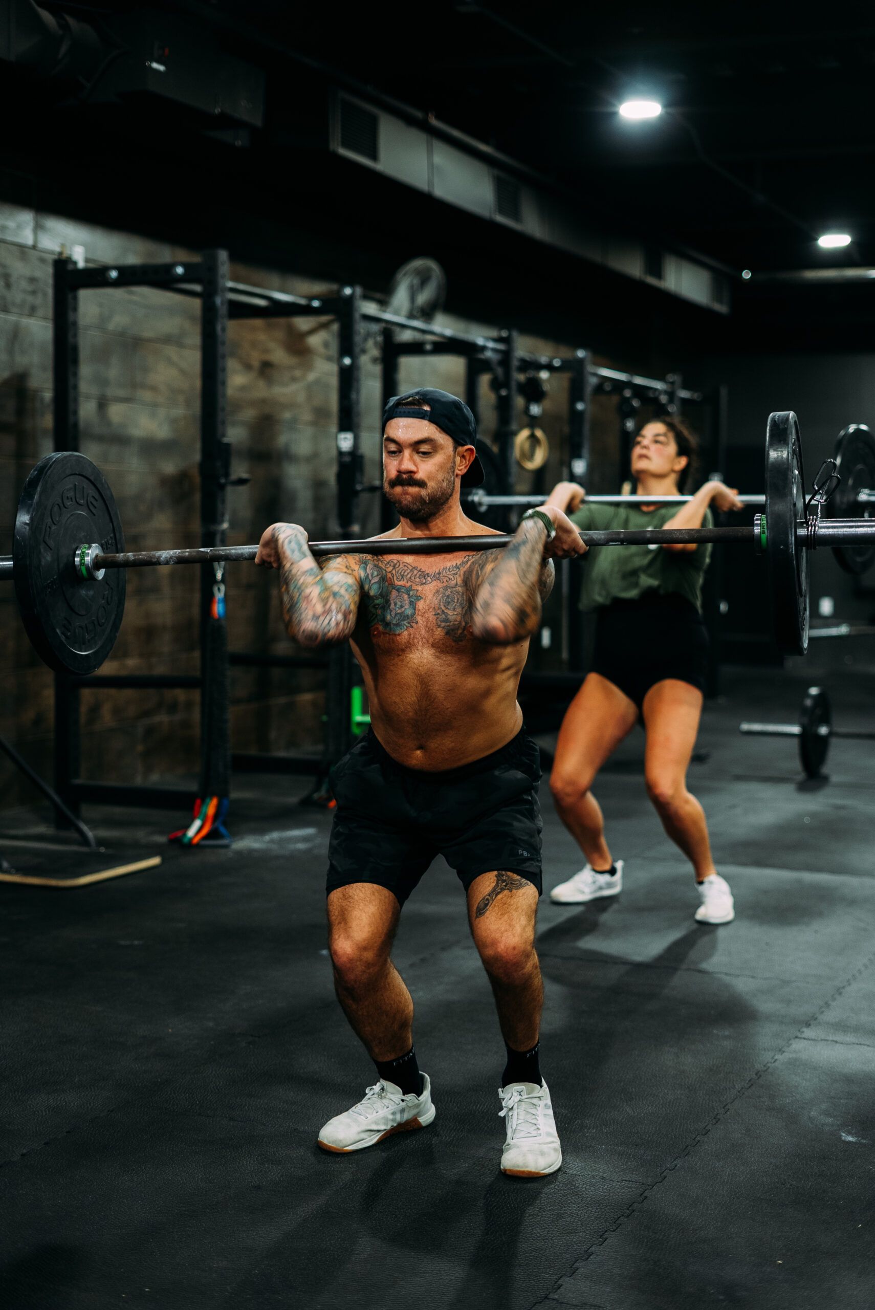 Two people lifting barbells in a dim gym, with a black rope and weight plates nearby.