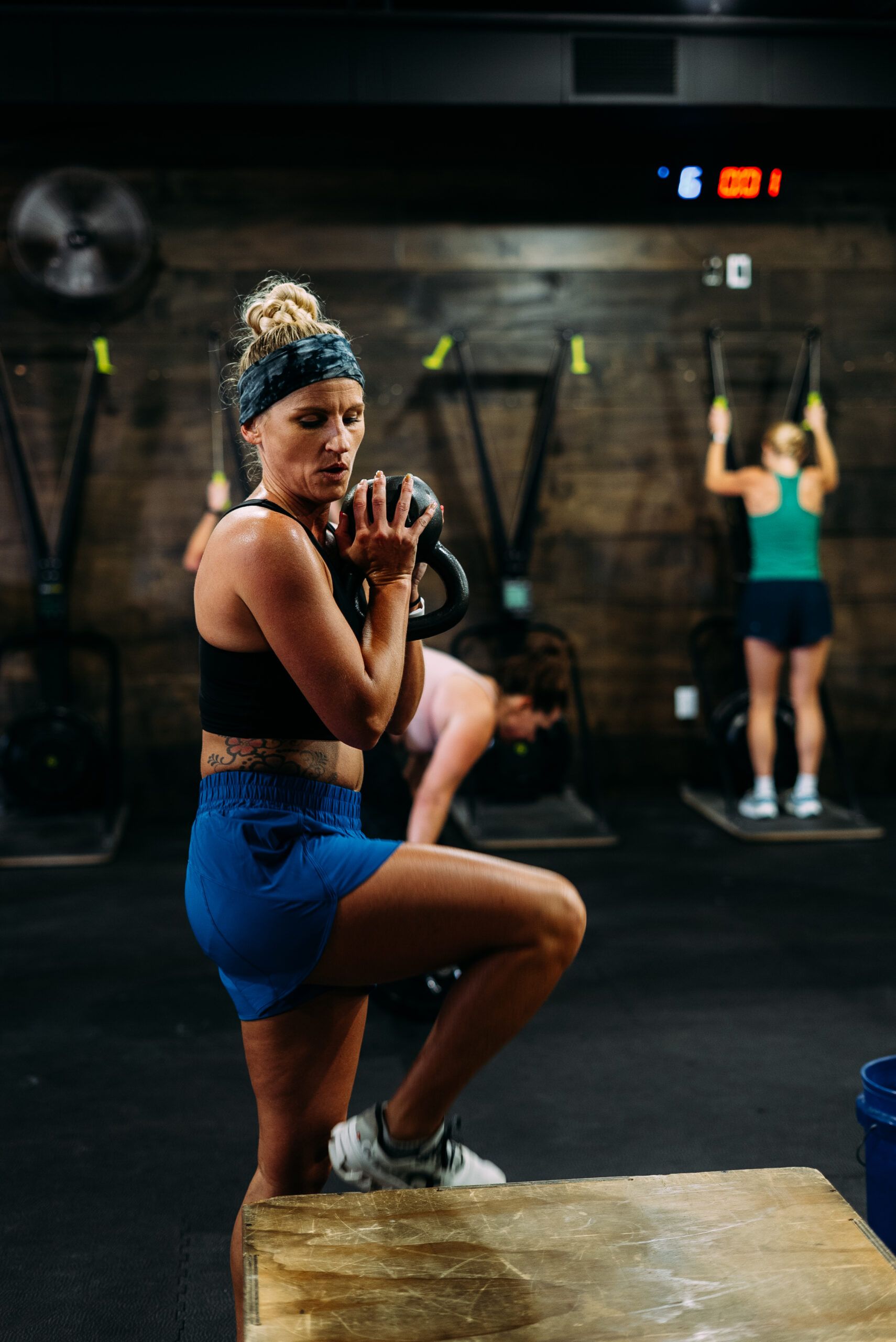 Woman exercising in a gym, stepping onto a box while holding a kettlebell, with another person in the background