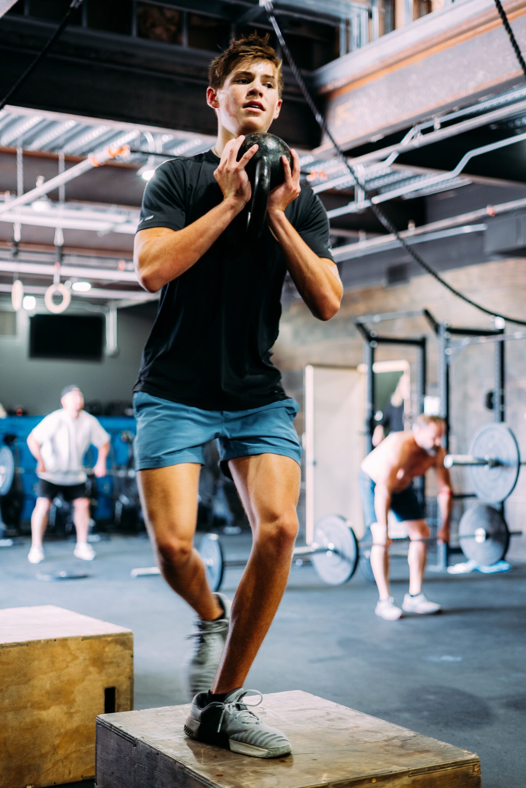 Person balancing on a wooden box in a gym while others exercise in the background