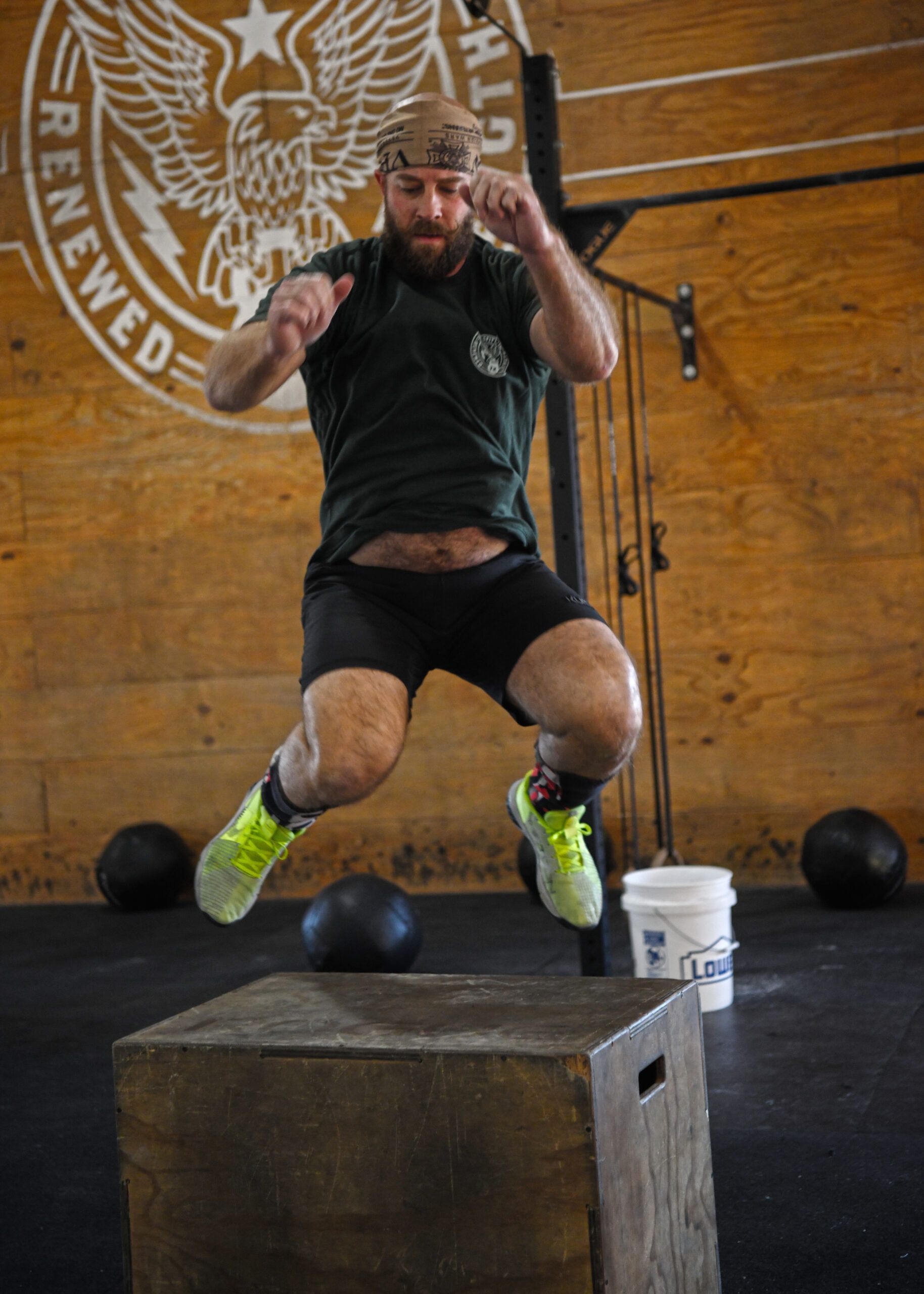 Athlete mid-jump onto a wooden box in a gym, with medicine balls and wooden walls behind him.