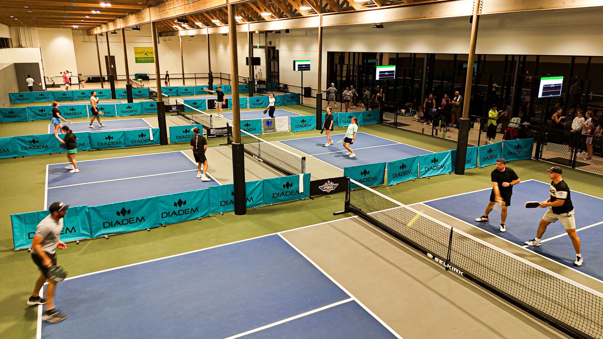 Indoor pickleball court with multiple players in a gym, wearing athletic gear and playing on blue courts.