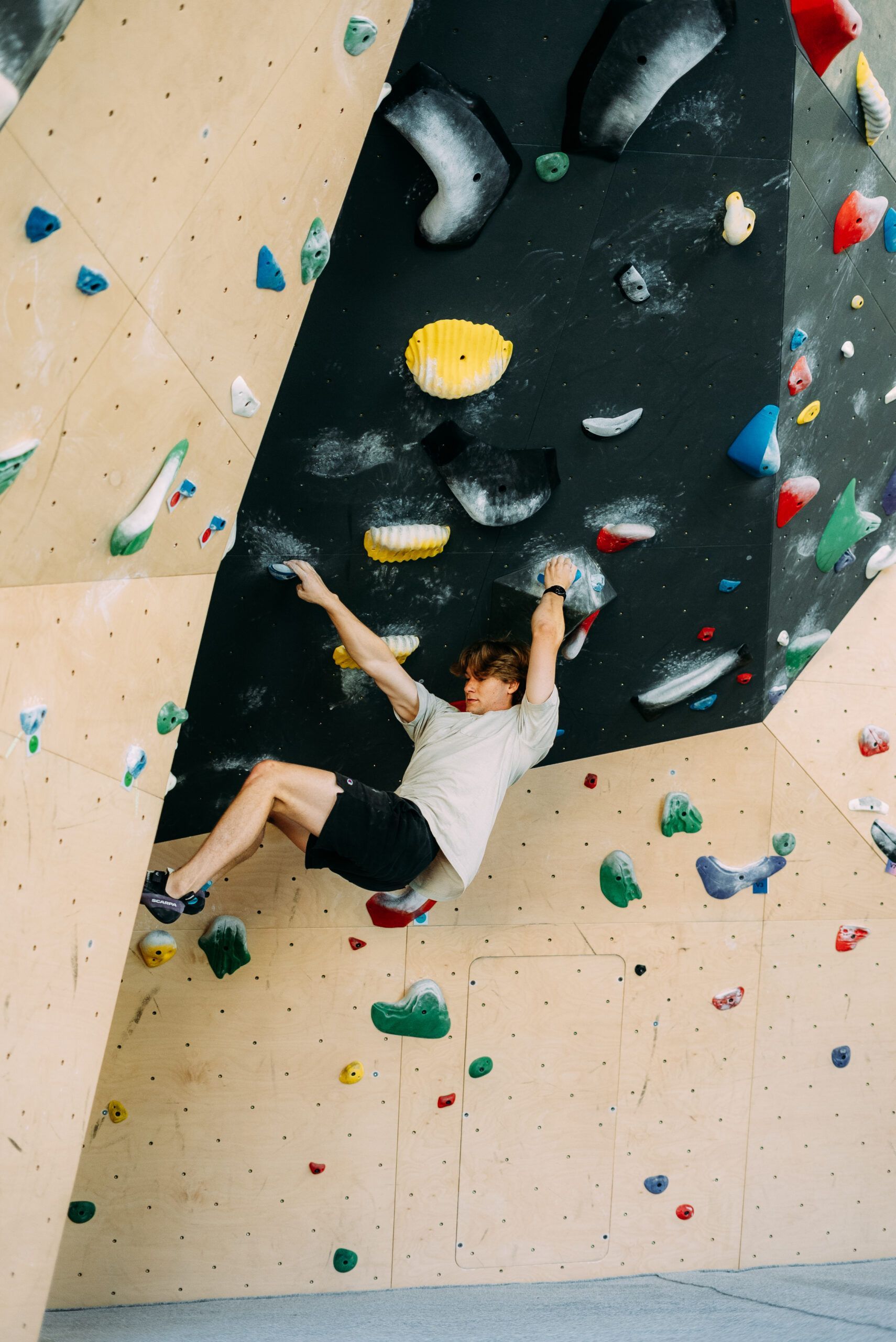 Person climbing an indoor rock wall with colorful holds, reaching upward beneath an overhang.