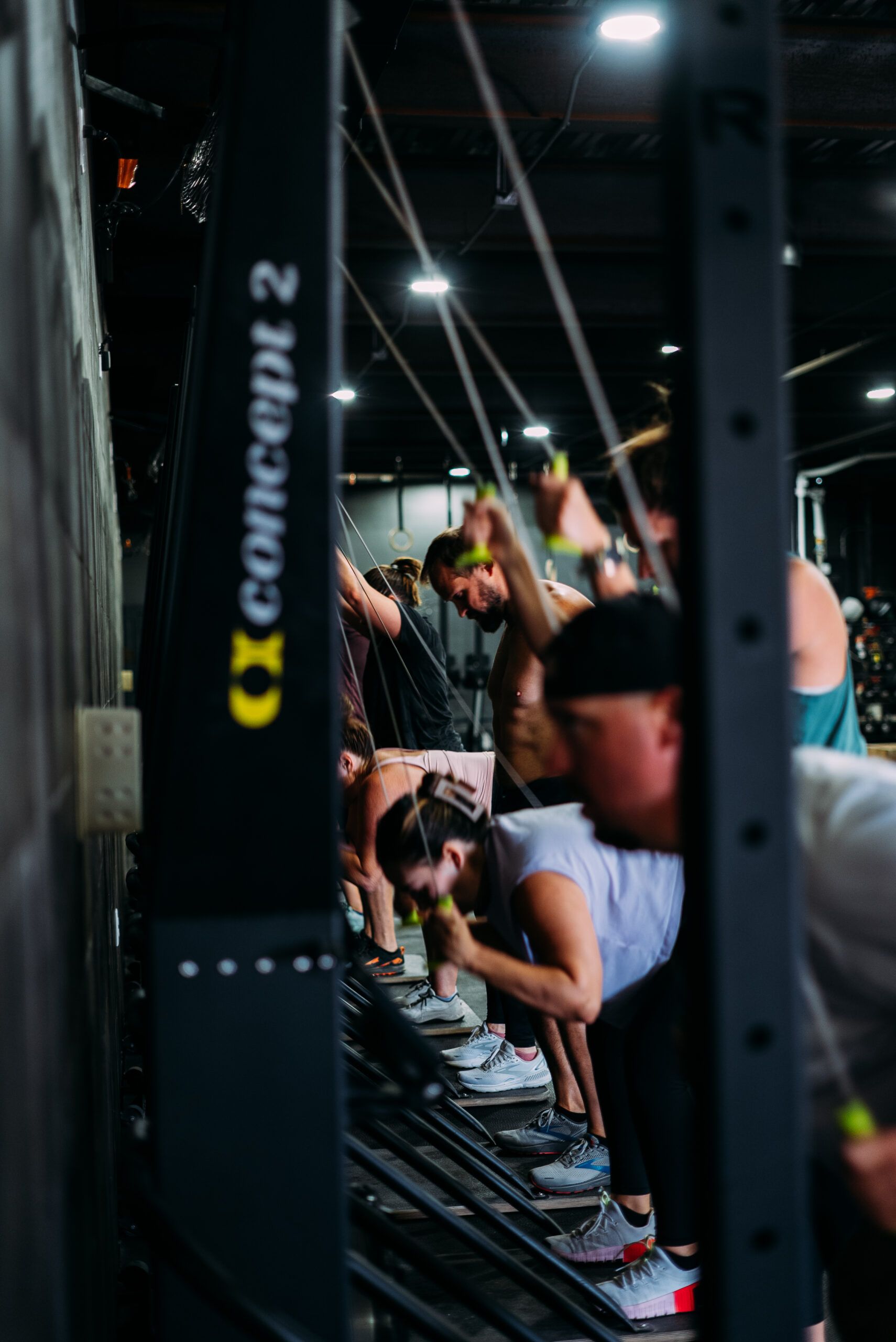 People using rowing machines in a dim gym, with a black Concept2 column in the foreground.