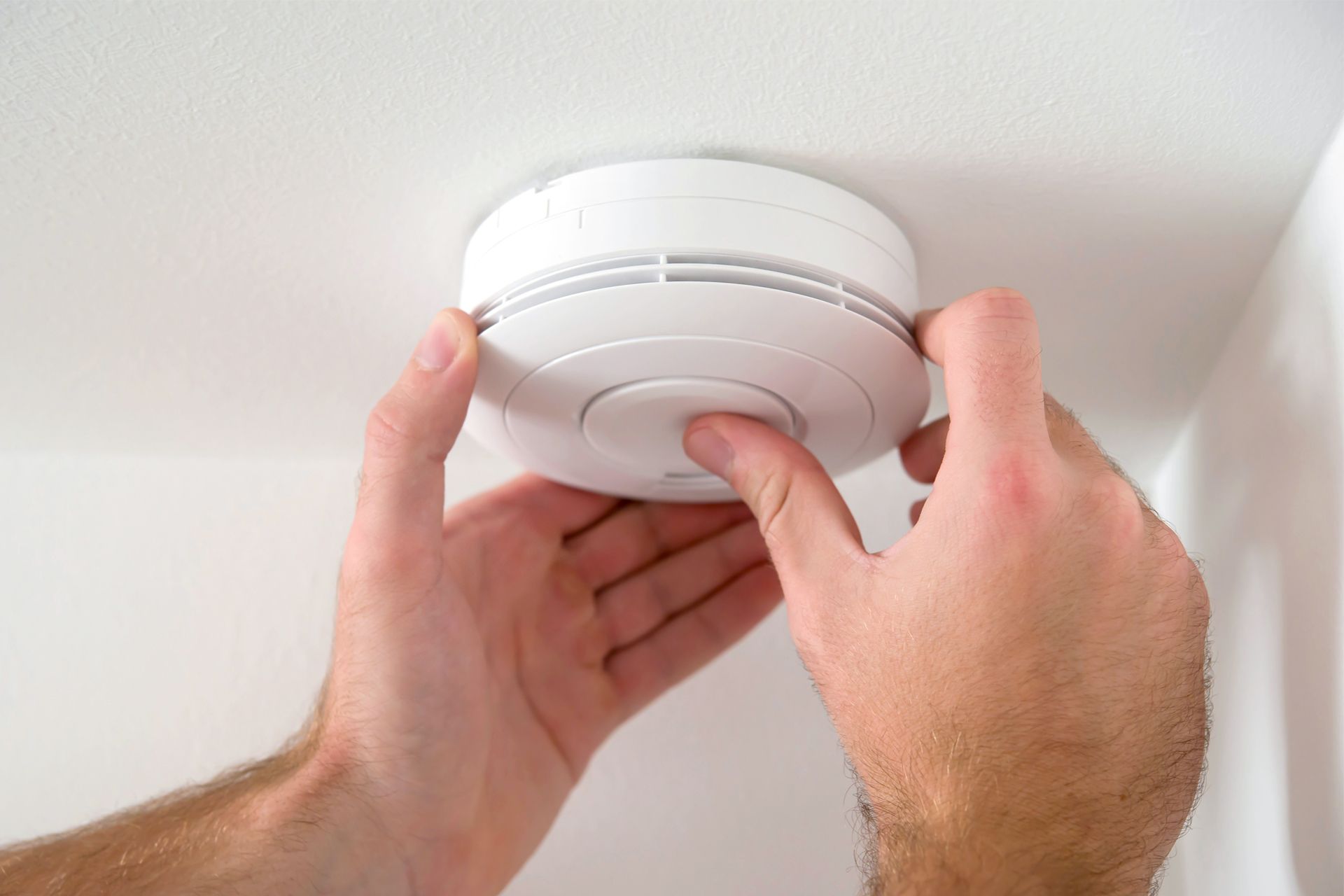 Hands installing a white smoke detector on a white ceiling.