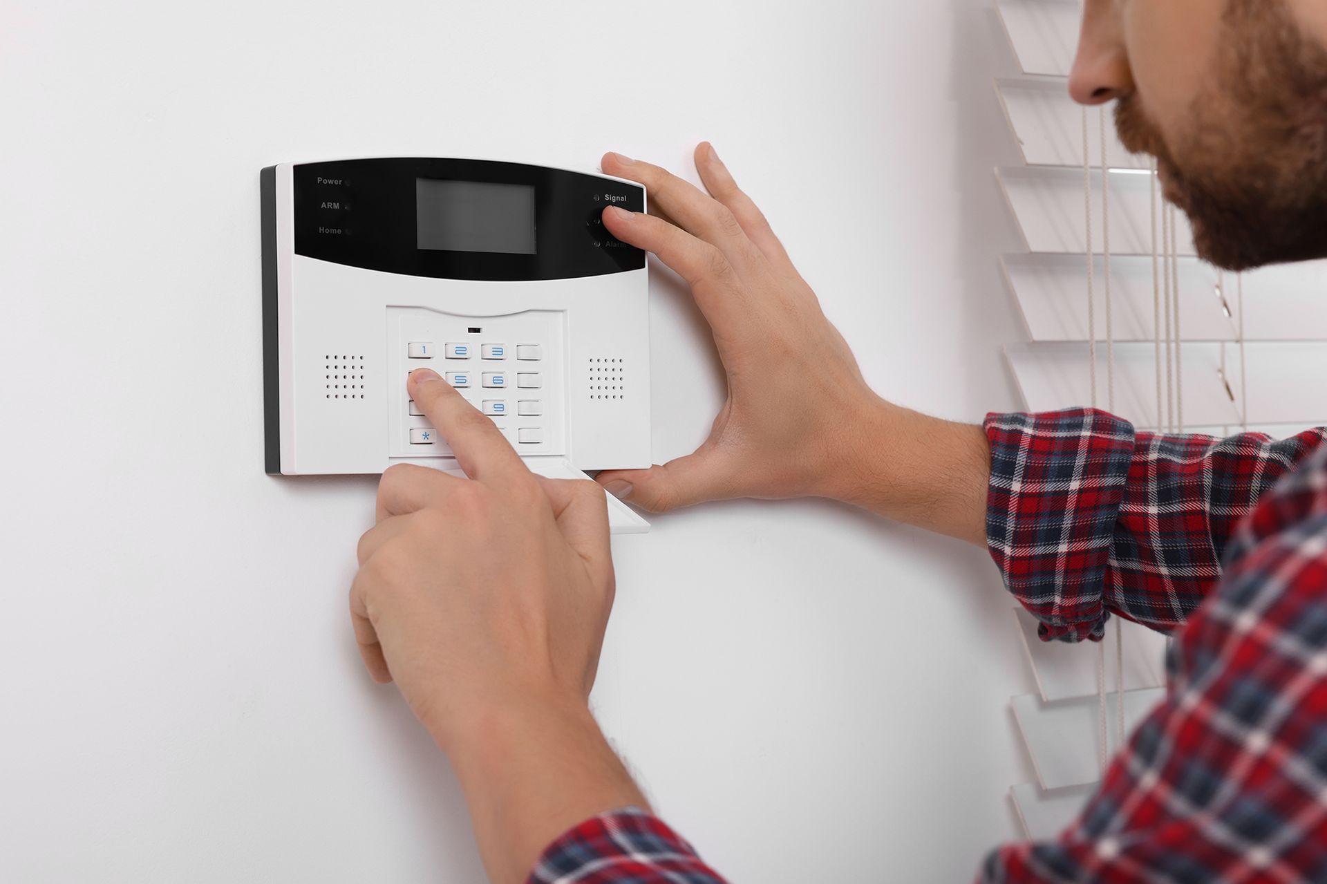 Man entering code on a white security system keypad on a wall, near blinds.