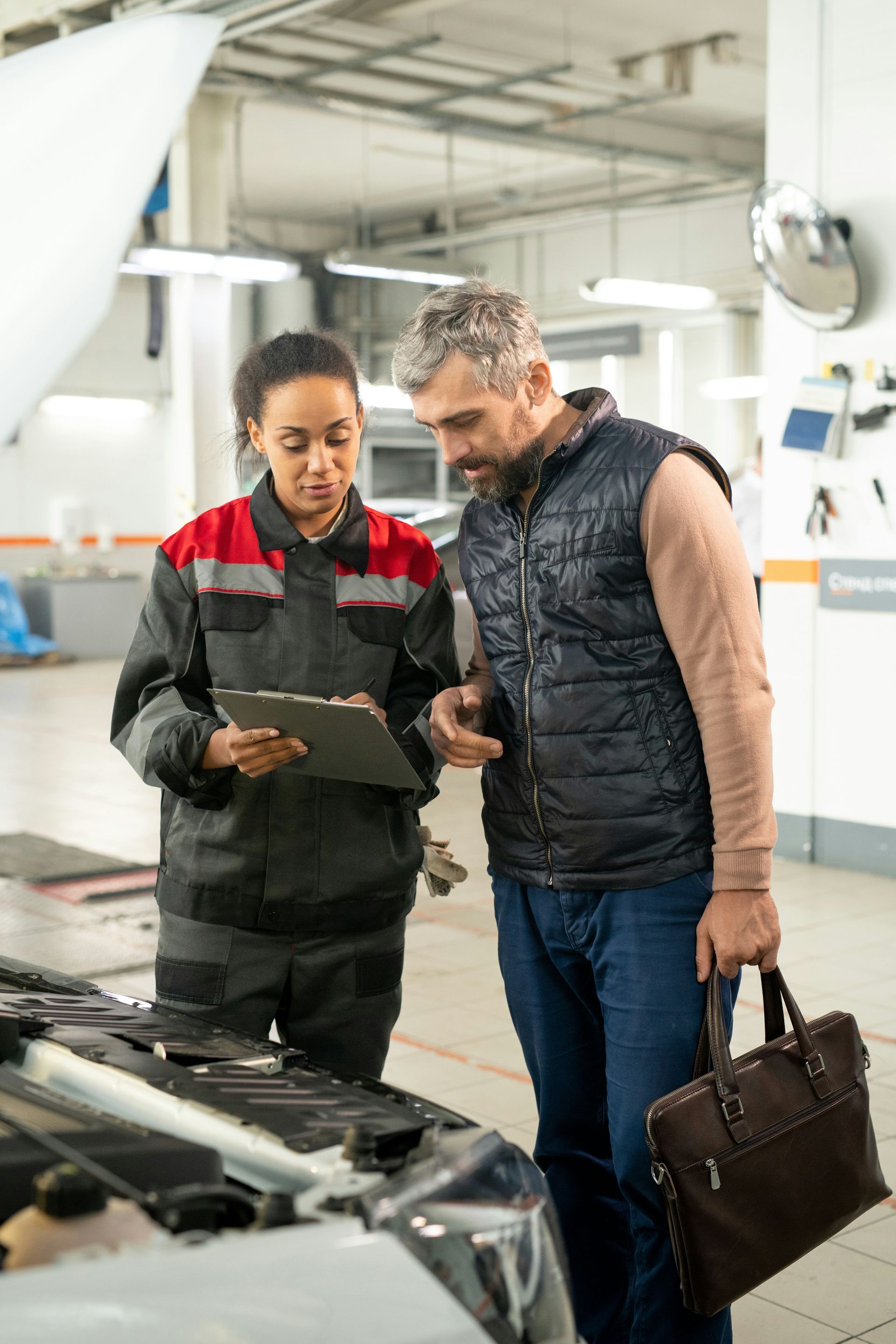 A man and a woman are looking under the hood of a car in a garage.