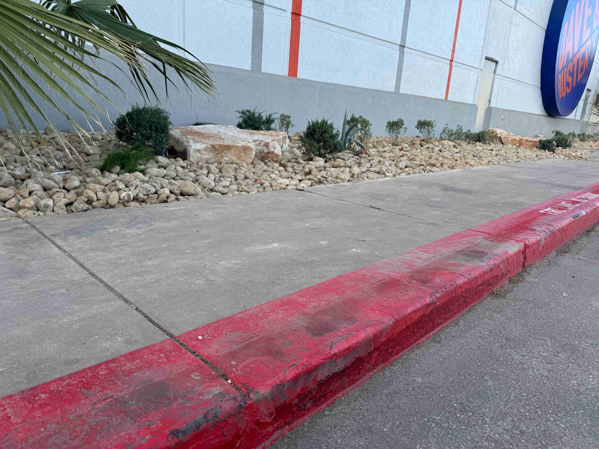 Red-painted curb along a sidewalk beside a building with decorative rocks and a palm plant.