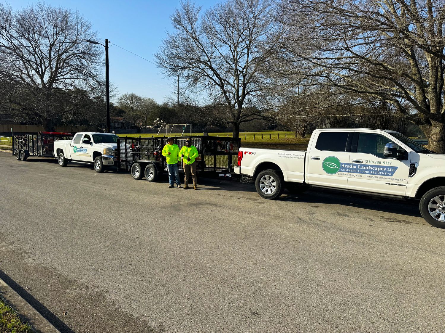 Two white trucks with trailers and workers wearing neon green stand on a road.