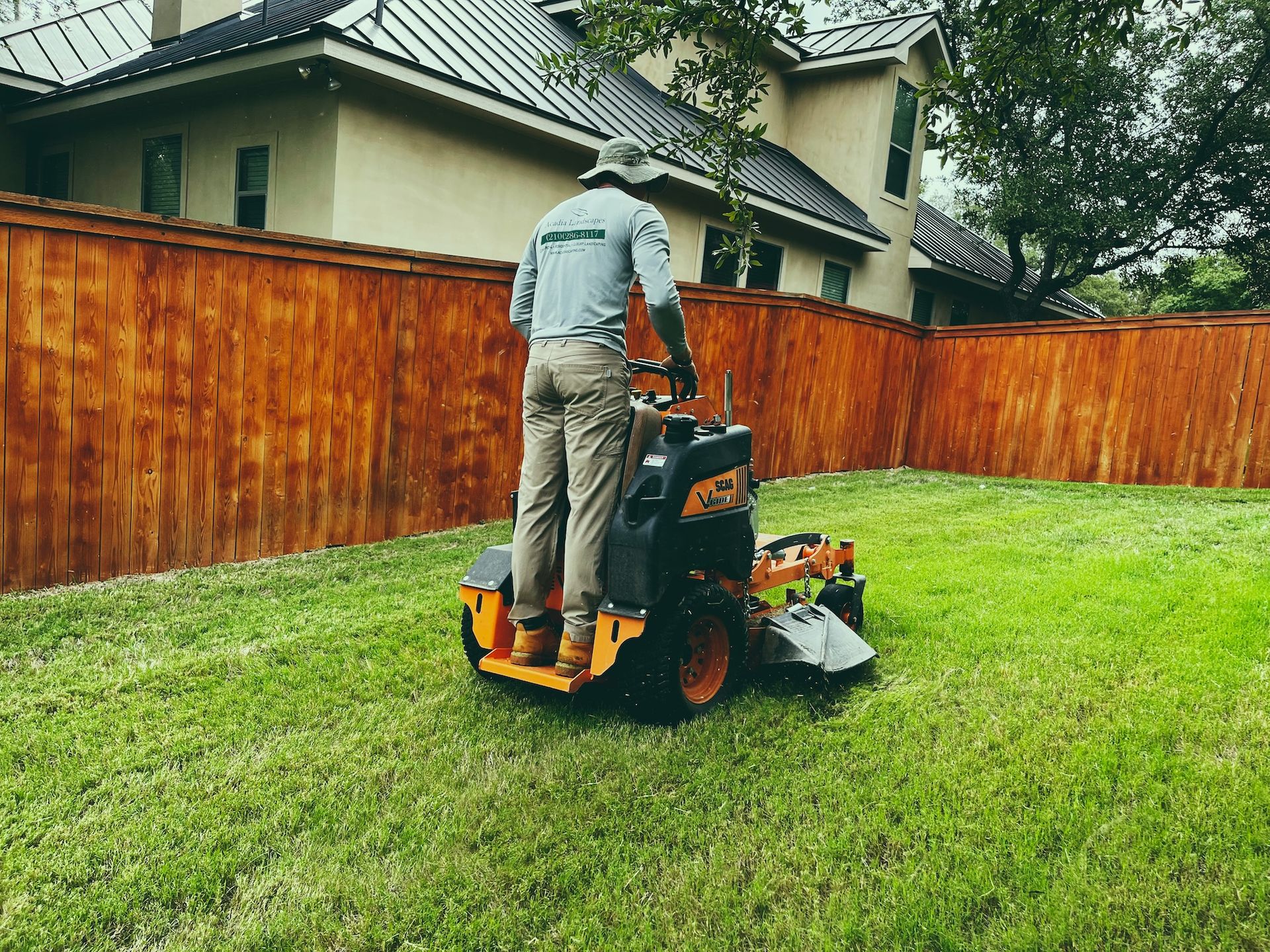 Man mowing a green lawn with a riding mower next to a stained wooden fence. A house is in the background.