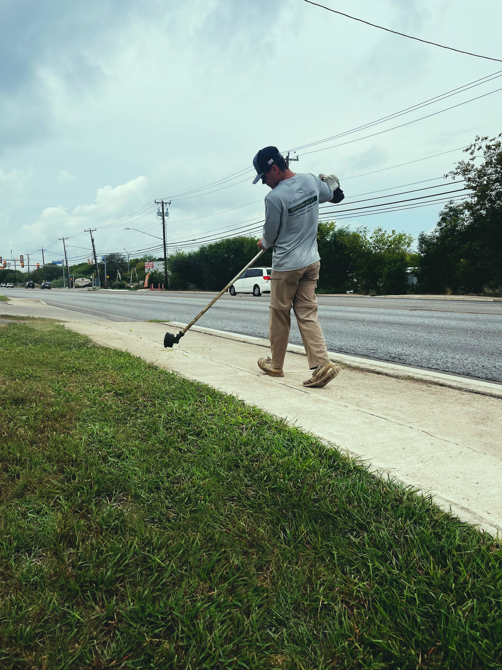 Man using a weed whacker along a sidewalk, grass in foreground, overcast sky.