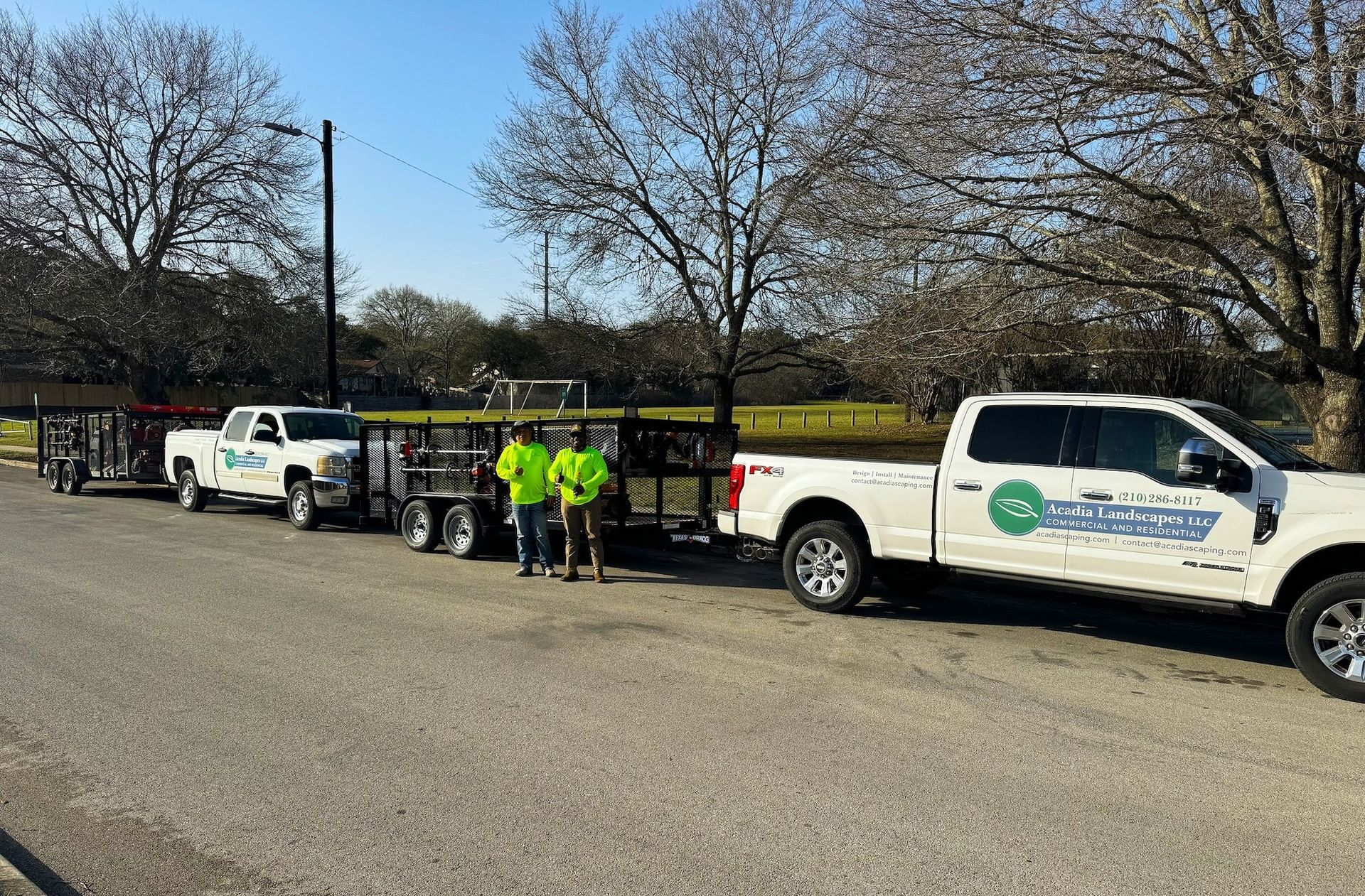 Two trucks with trailers and two workers wearing neon green near trees and a field on a sunny day.
