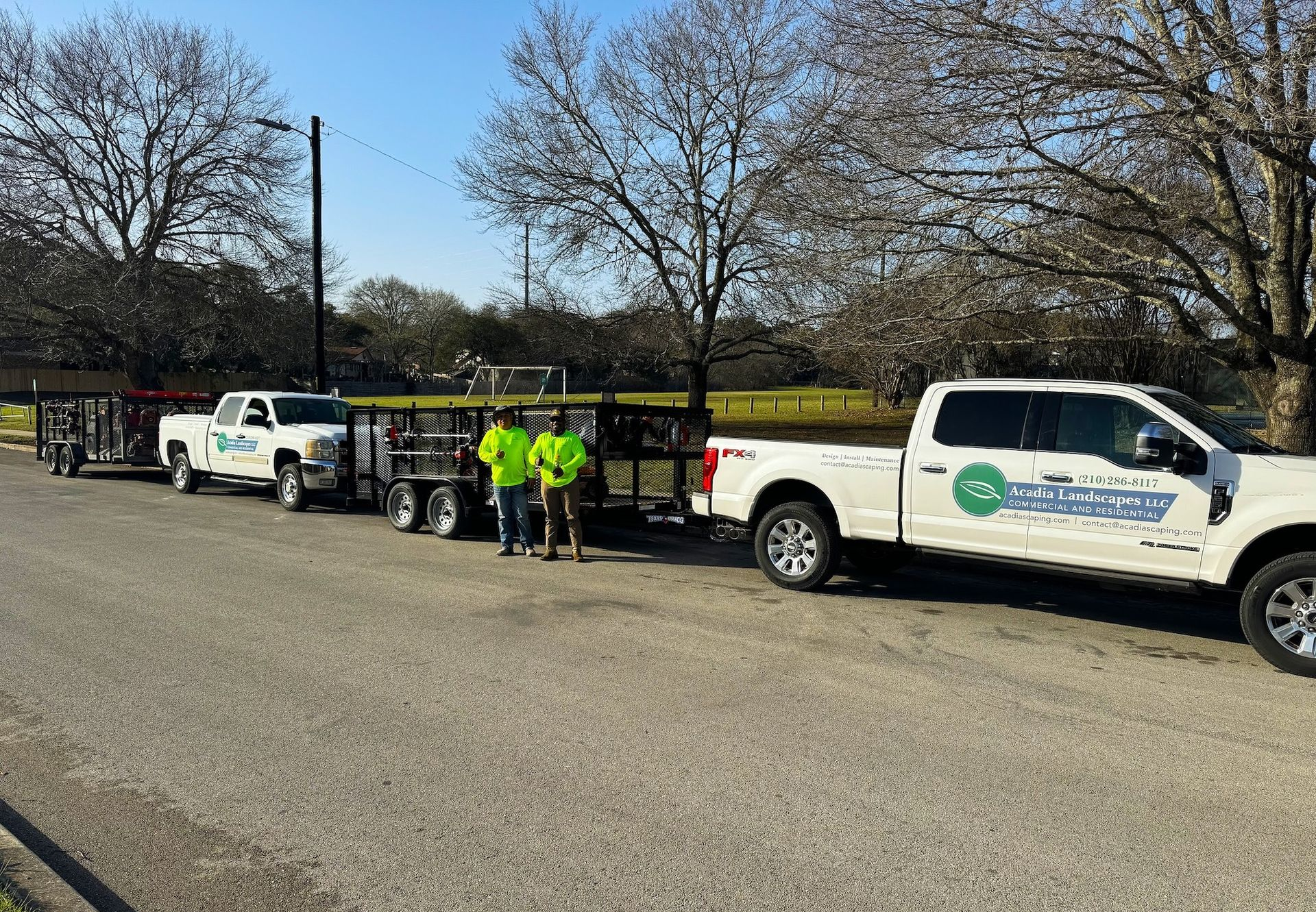 Two trucks with trailers and two people in high-vis vests parked on road; trees and field in background.