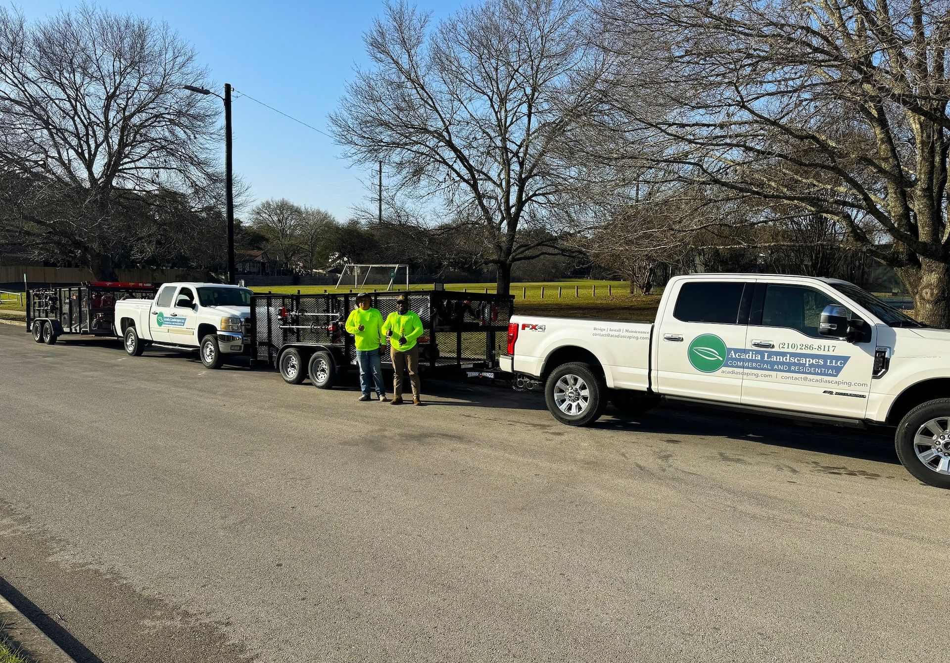 Two white trucks with trailers parked on a paved lot, two workers stand nearby in green. Trees in background.