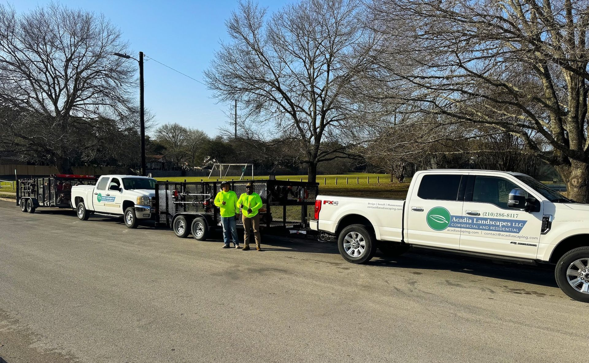 Two white trucks with trailers parked, two people in green vests standing nearby.