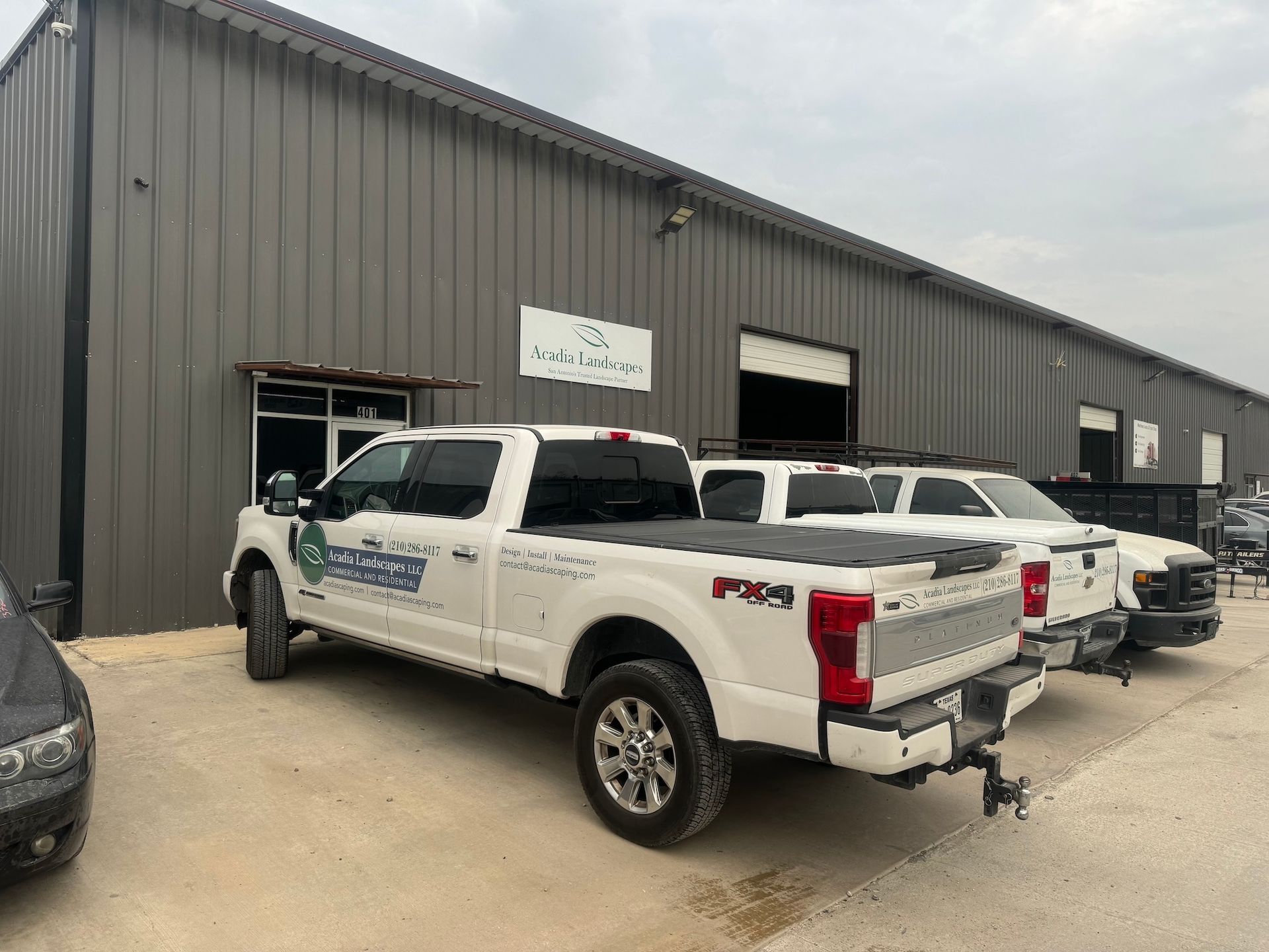 White pickup trucks parked outside a gray commercial building; sign above door.