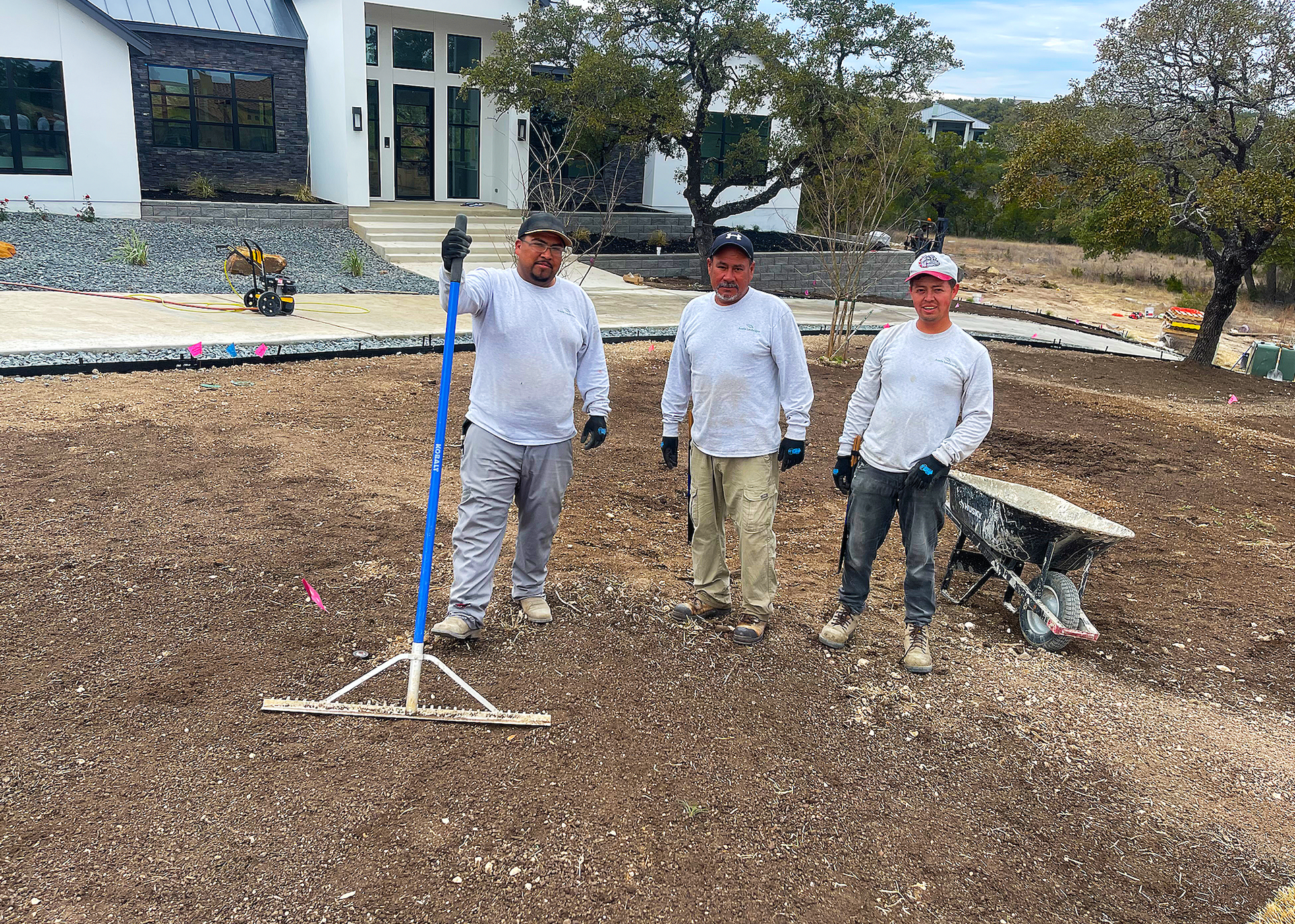 Three workers standing on a gravel bed, in front of a house. One holds a rake.