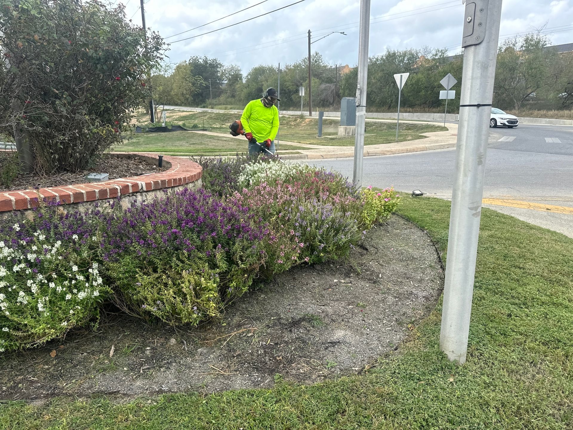 Person in neon vest trims bushes with a trimmer near a road with a sign and car.