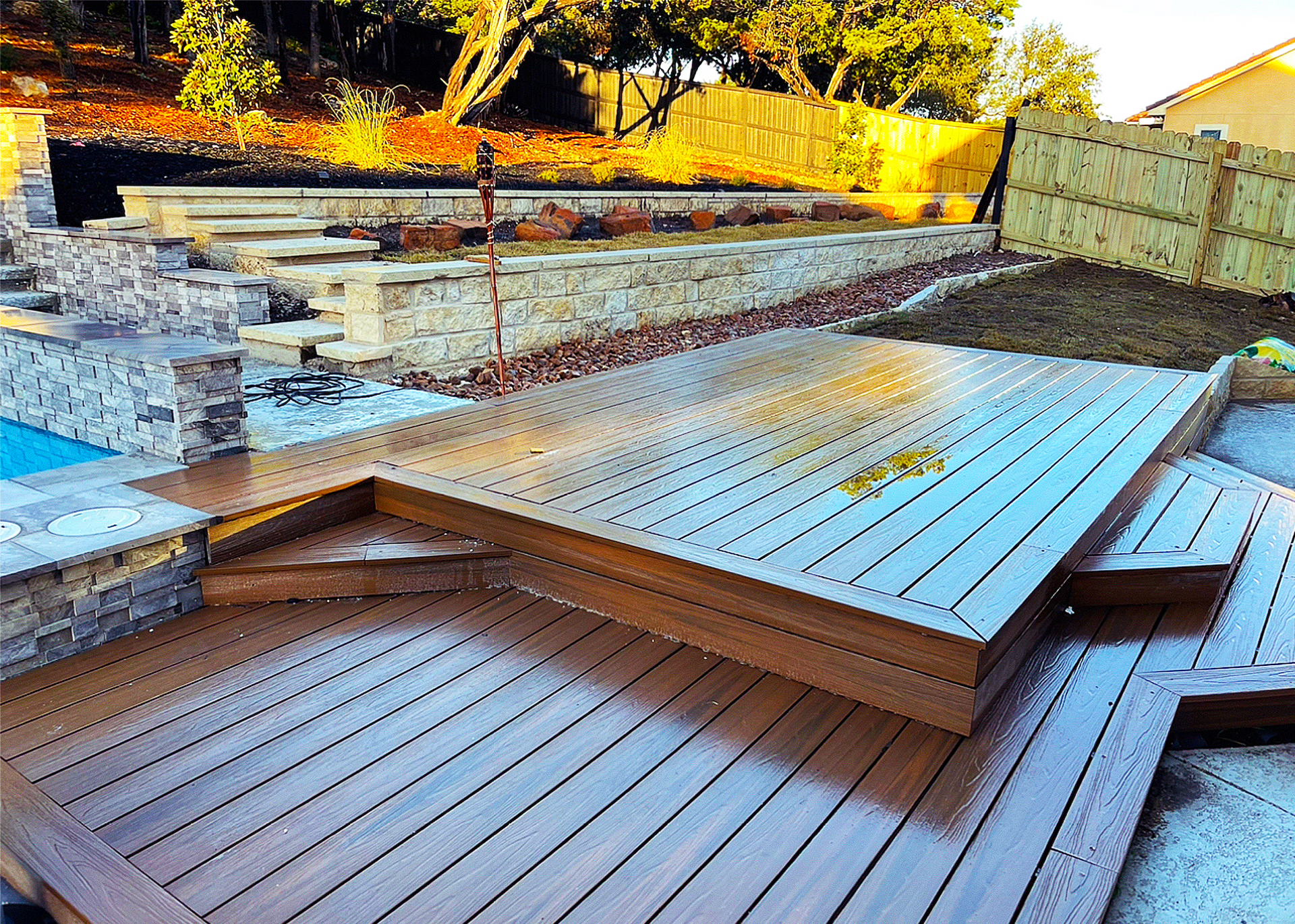 Wooden deck with tiered landscaping, pool visible to the left, and a fence in the background.