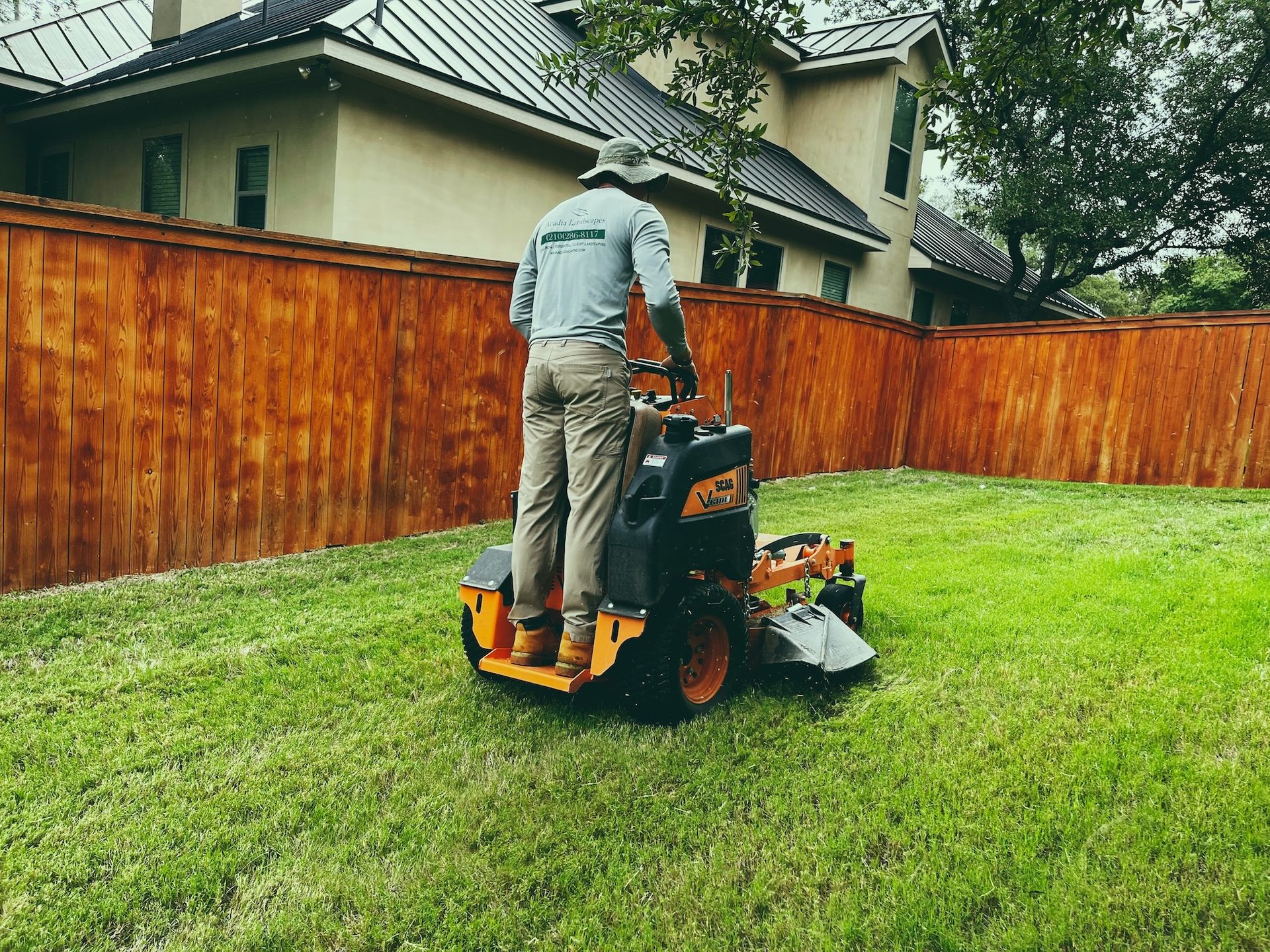 Person mowing a green lawn with a riding mower next to a brown wooden fence and a house.
