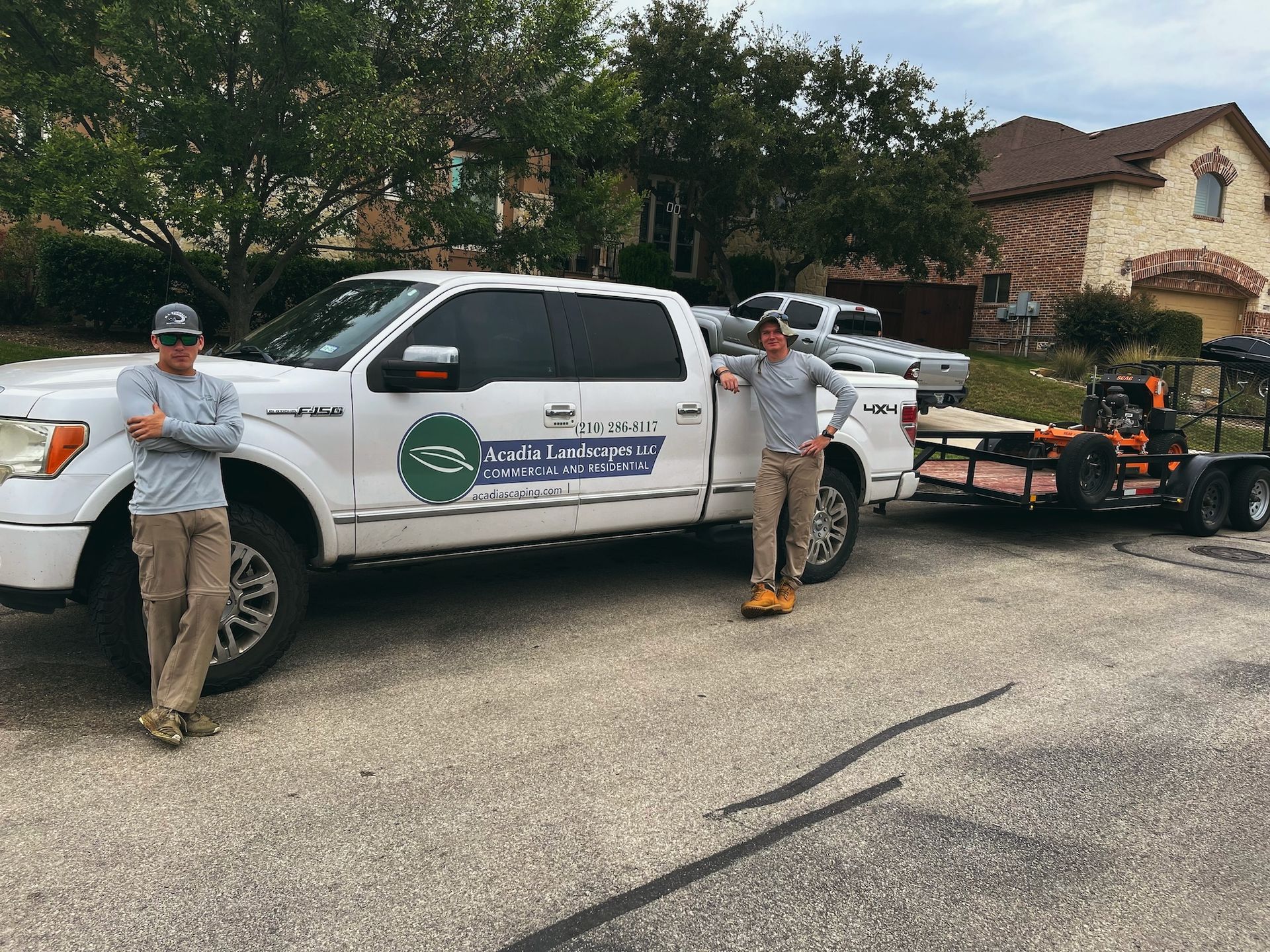 Two men stand by a white pickup truck with a landscaping business logo, a trailer attached, in front of a house.