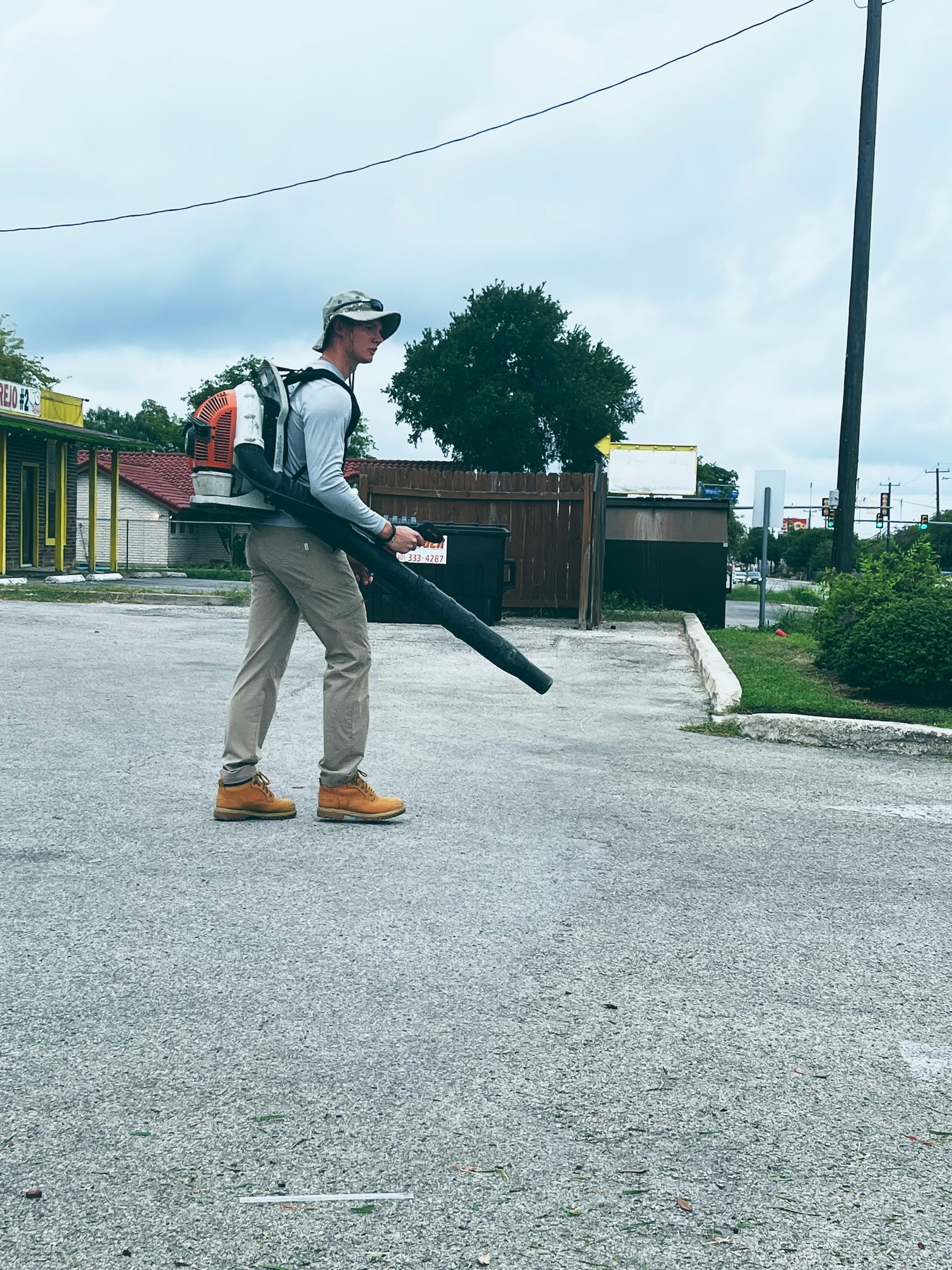 Man using a backpack leaf blower on a paved surface. Cloudy sky overhead.