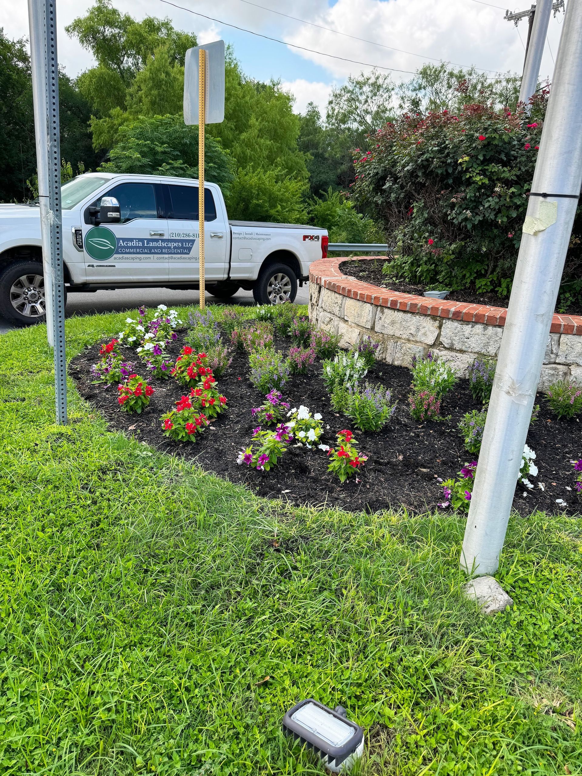 Flowerbed with a truck in front, trees, and sign. Red, white, and purple flowers in dark mulch. Green grass in the foreground.