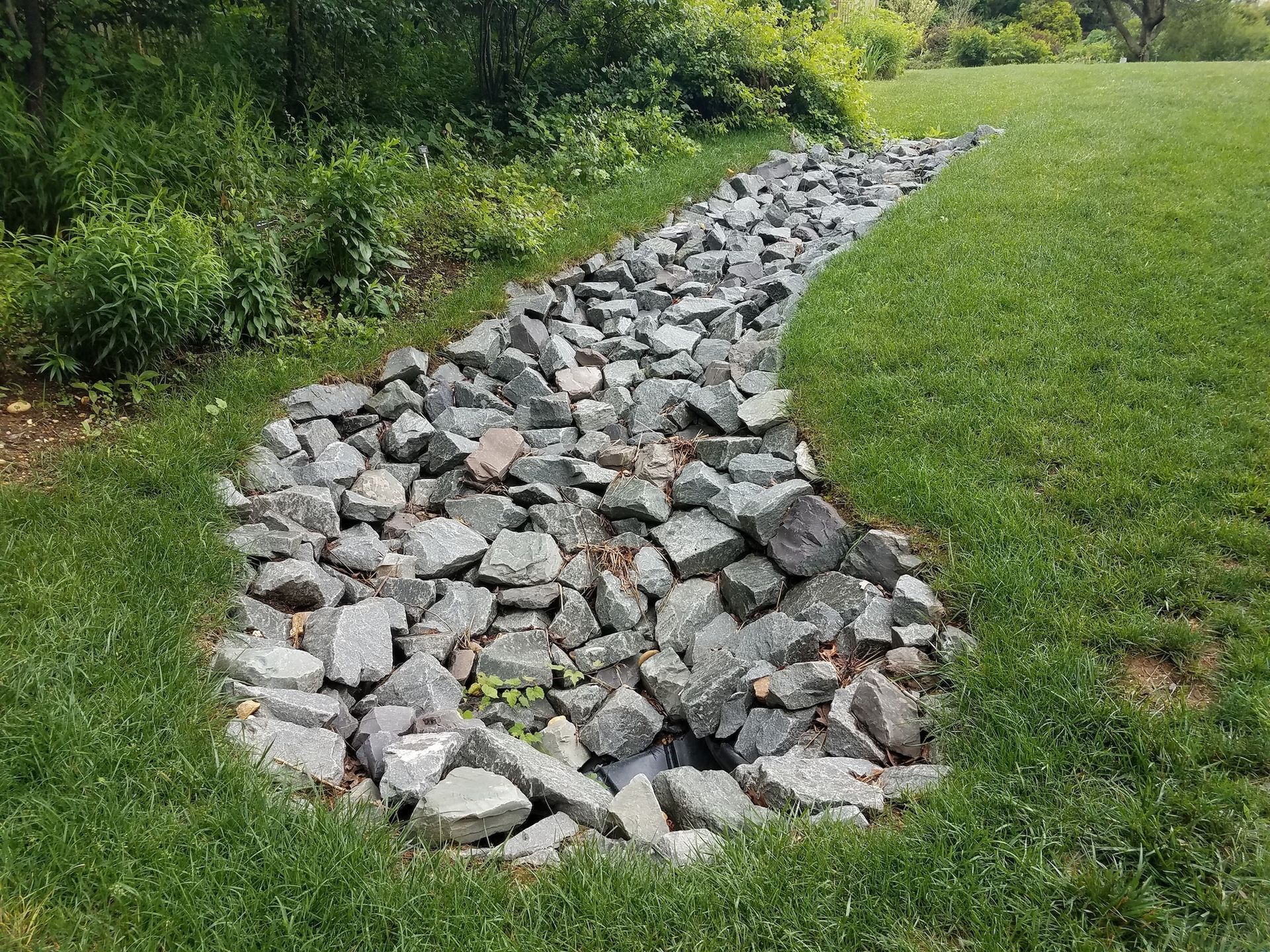 Rock-lined drainage channel running through a grassy yard beside shrubs and trees