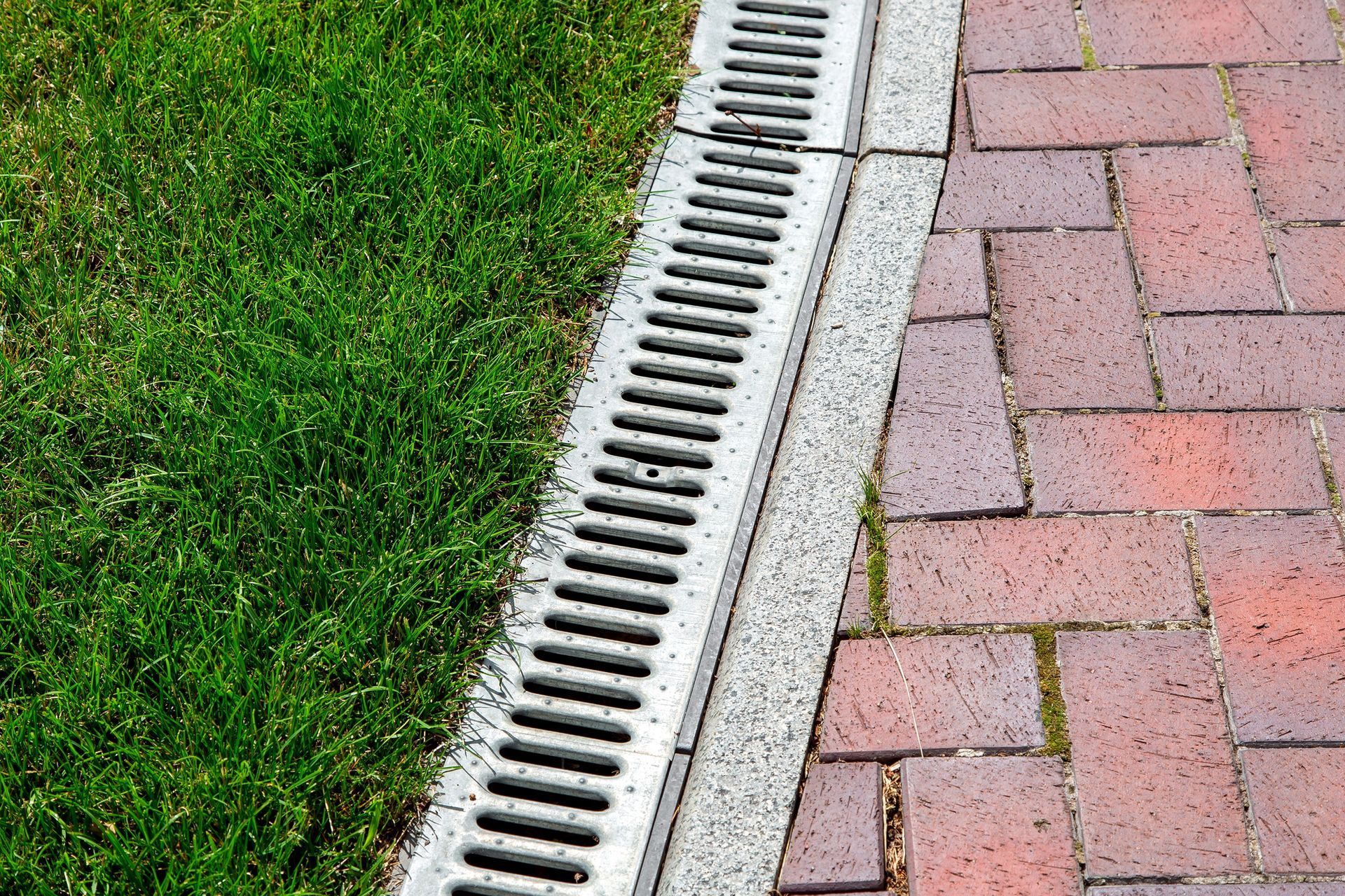 Grass beside a silver drainage grate and red brick sidewalk